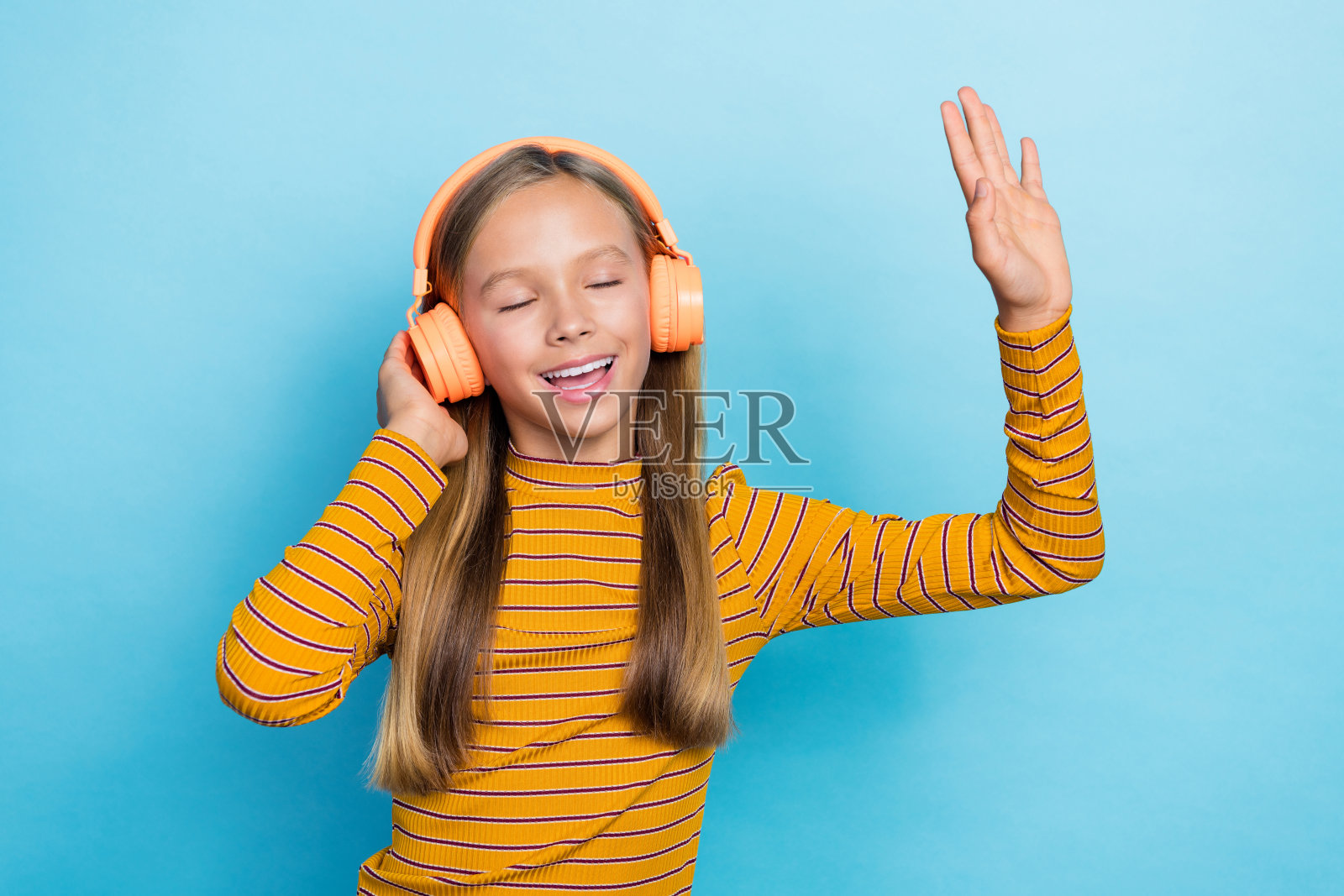 兴奋无忧的女孩闭着眼睛,手触碰耳机,享受旋律,背景为蓝色。照片摄影图片