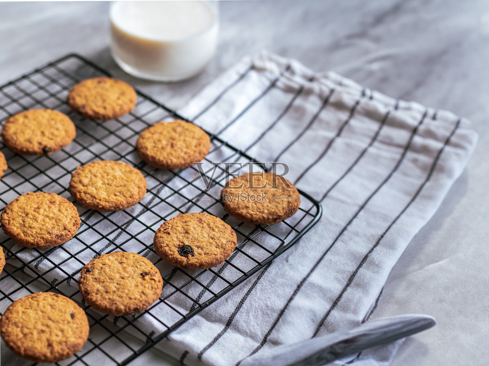 新鲜出炉的燕麦饼干在黑色铁架上冷却,背景明亮,柔和的自然光照射。饼干呈金棕色,带有葡萄干的点缀,口感酥脆,营造出温馨的氛围。照片摄影图片