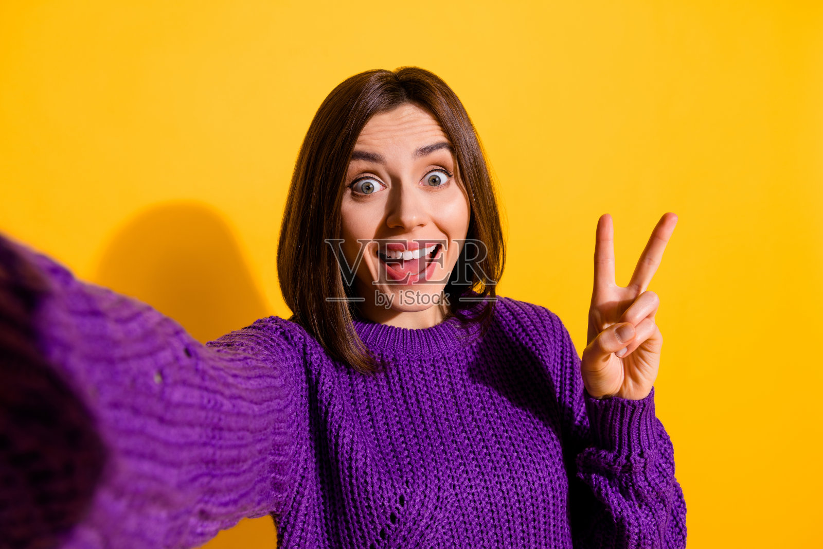 迷人的惊讶女孩自拍,做出V字手势,背景为黄色。照片摄影图片