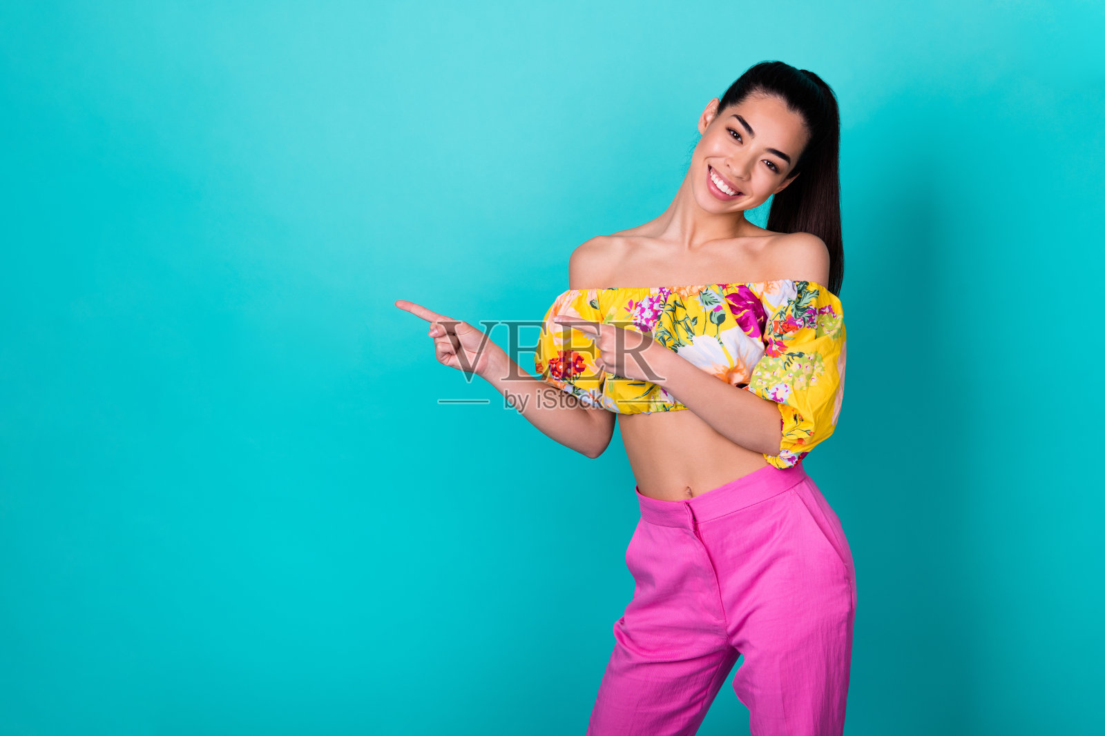 漂亮的千禧女性穿着花卉上衣和粉色裤子,独自站在青色背景前。照片摄影图片