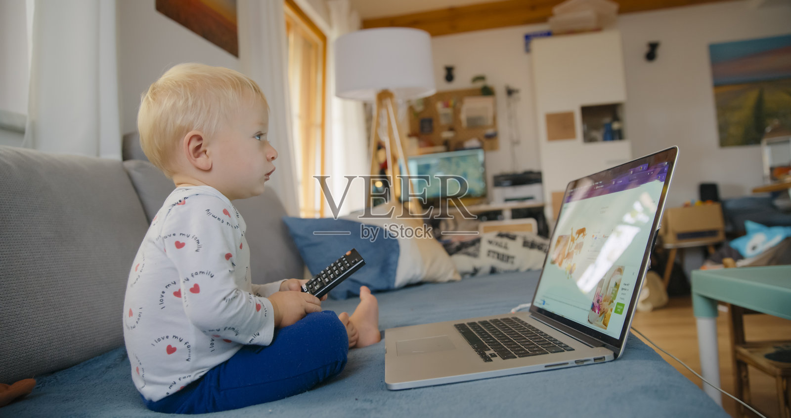 坐在沙发上的小男孩,手里拿着笔记本电脑和电视遥控器照片摄影图片