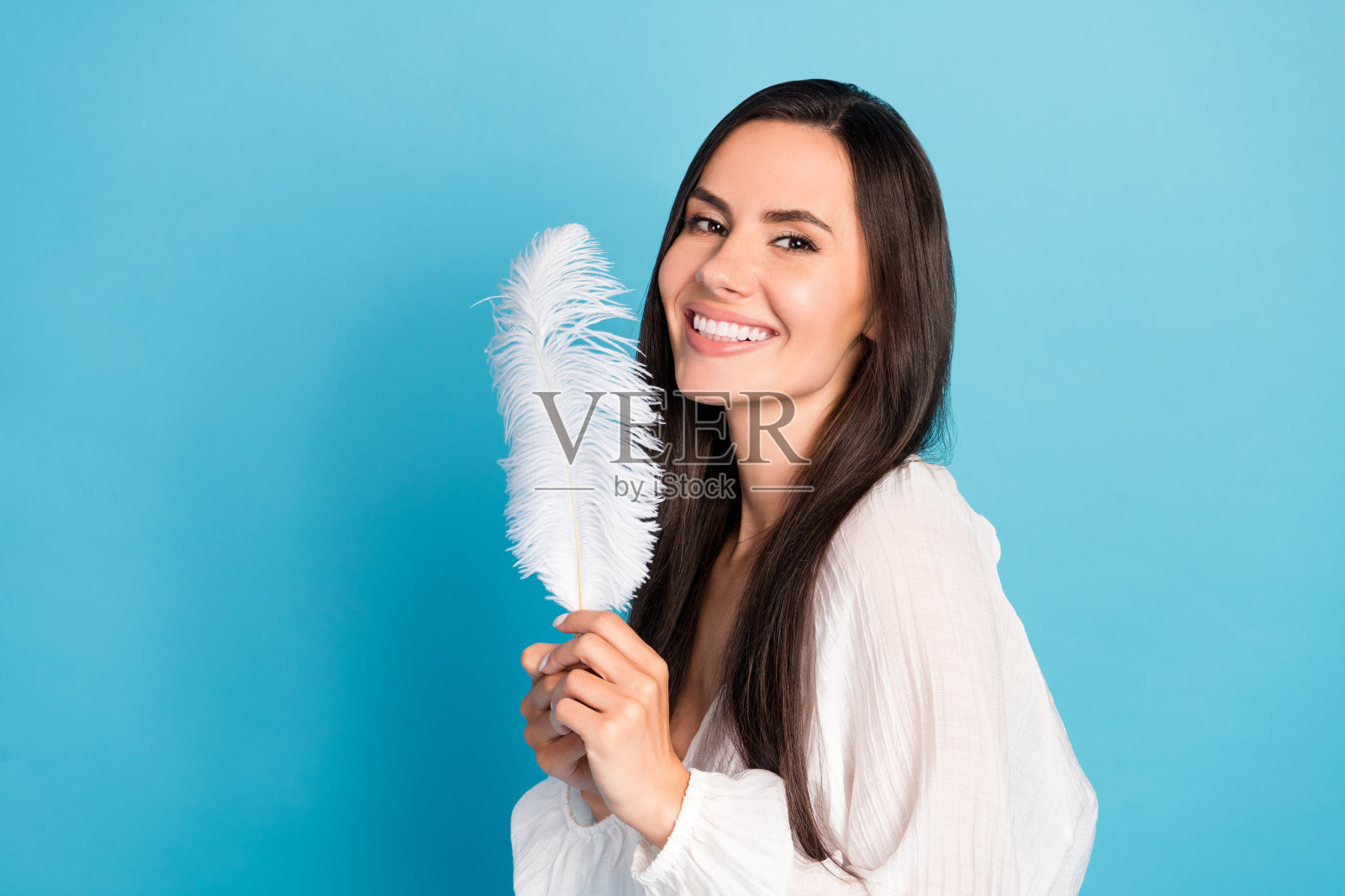 漂亮的棕发女士手持羽毛,穿着白色衬衫,背景为蓝色。照片摄影图片