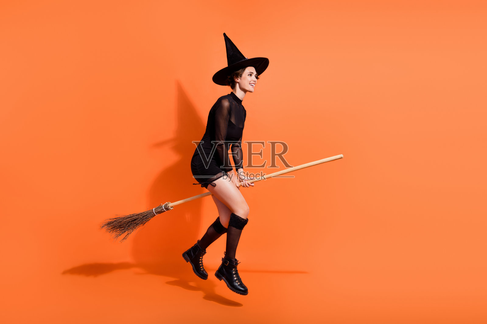 全身照:漂亮的女人骑着扫帚,穿着时尚的黑色万圣节服装,独自在橙色背景上购物。照片摄影图片