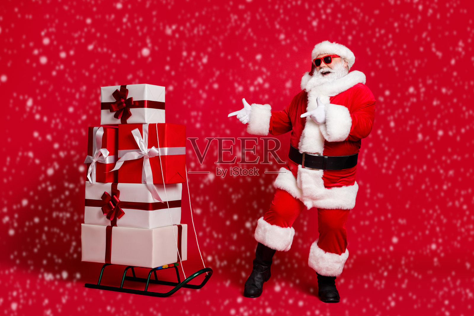 全身照的退休老人,白色胡须,手指指向雪橇,旁边有许多礼品盒,穿着圣诞老人服装,戴着太阳镜和帽子,脚穿黑靴,背景为红色。照片摄影图片