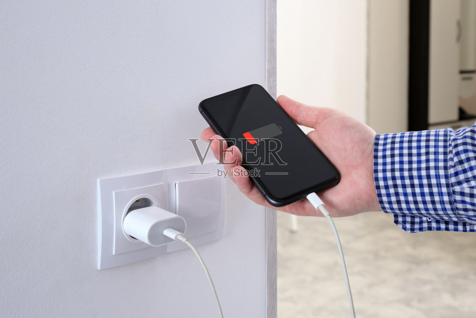 一个男人手里拿着智能手机,连接着电源插座的充电线。背景是一个住宅公寓。照片摄影图片
