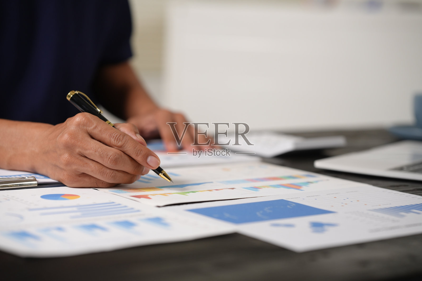 一位专注的商人在办公室桌前坐着,分析纸质图表,同时使用笔记本电脑。他正在审查财务和市场报告,专注于数据以进行战略决策。照片摄影图片