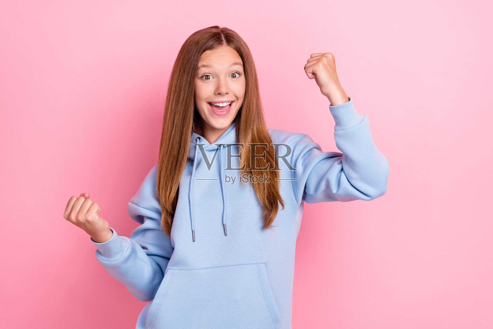 年轻漂亮可爱的女孩学生穿着蓝色连帽衫,双拳举起庆祝,兴奋地表现出胜利的姿态,背景为粉色。照片摄影图片