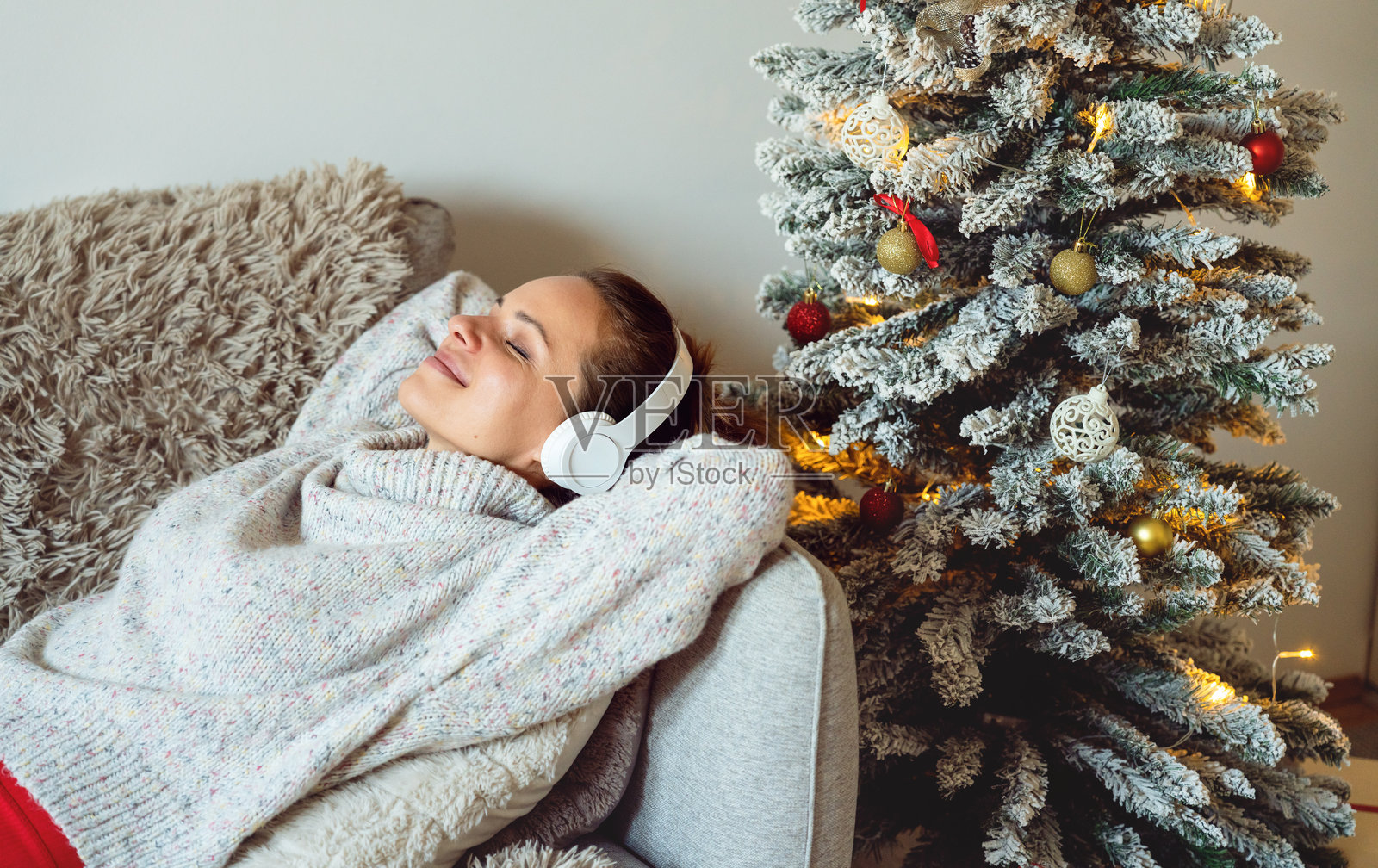 女人戴着无线耳机躺在沙发上,旁边是圣诞树,正在听轻松的音乐。照片摄影图片