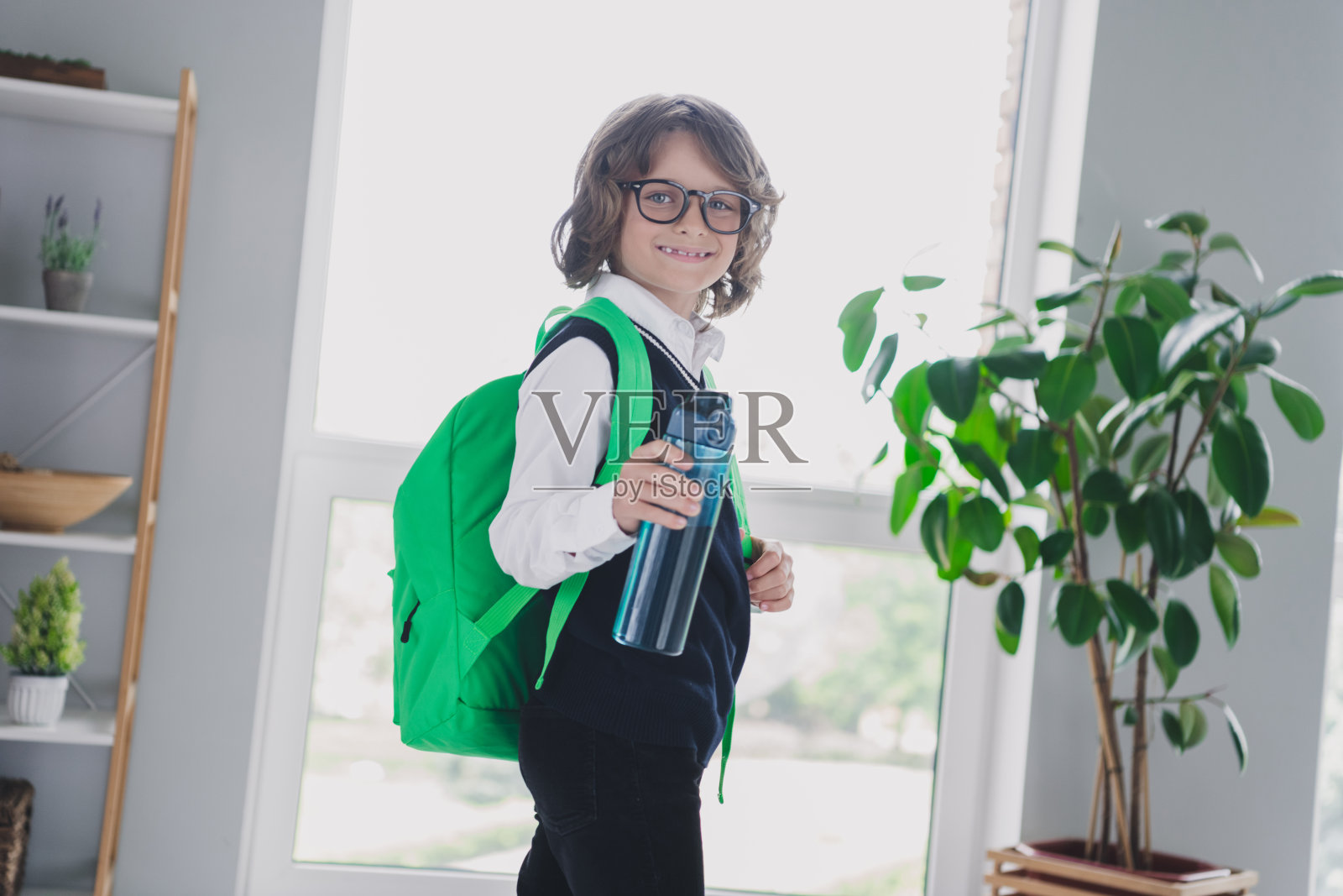 可爱的幽默聪明男孩在家中站着,准备上学,手里拿着水,室内环境。照片摄影图片