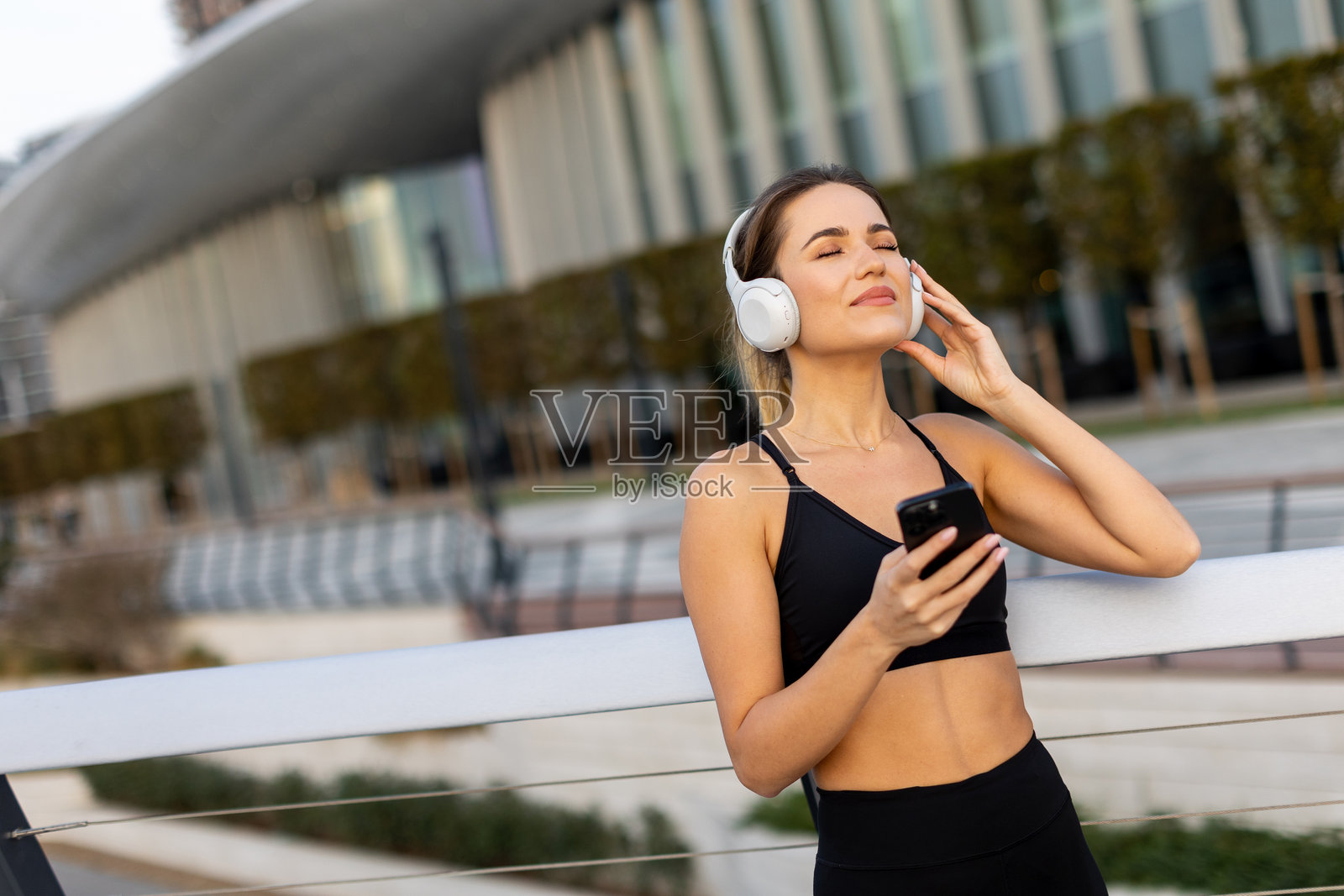 女人在城市公园的现代建筑外享受阳光明媚的日子,边锻炼边听音乐。照片摄影图片