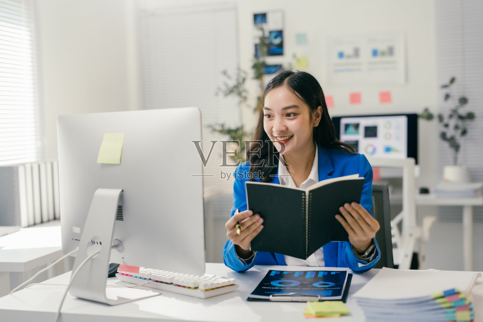 年轻的亚洲女商人穿着蓝色西装,微笑着阅读笔记,同时在明亮的现代办公室里通过电脑进行视频通话。照片摄影图片