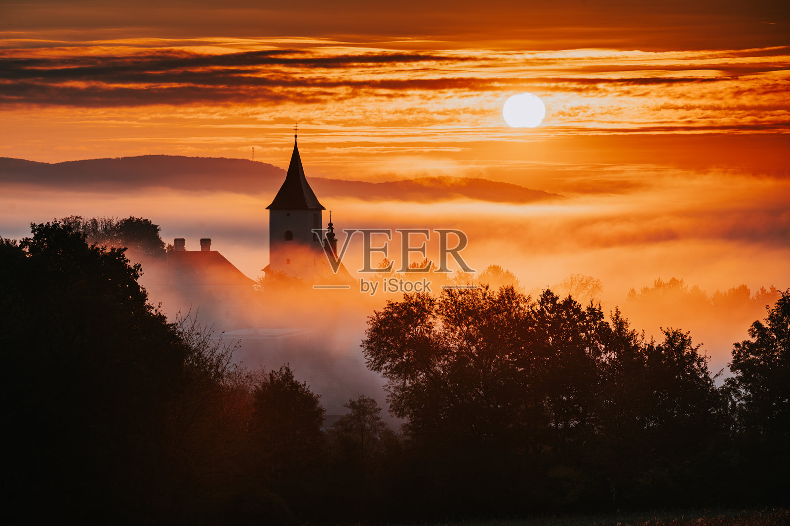 美丽的秋晨日出时基督教教堂的剪影,乡村风景中橙色的天空和柔和的晨雾笼罩着村庄。照片摄影图片