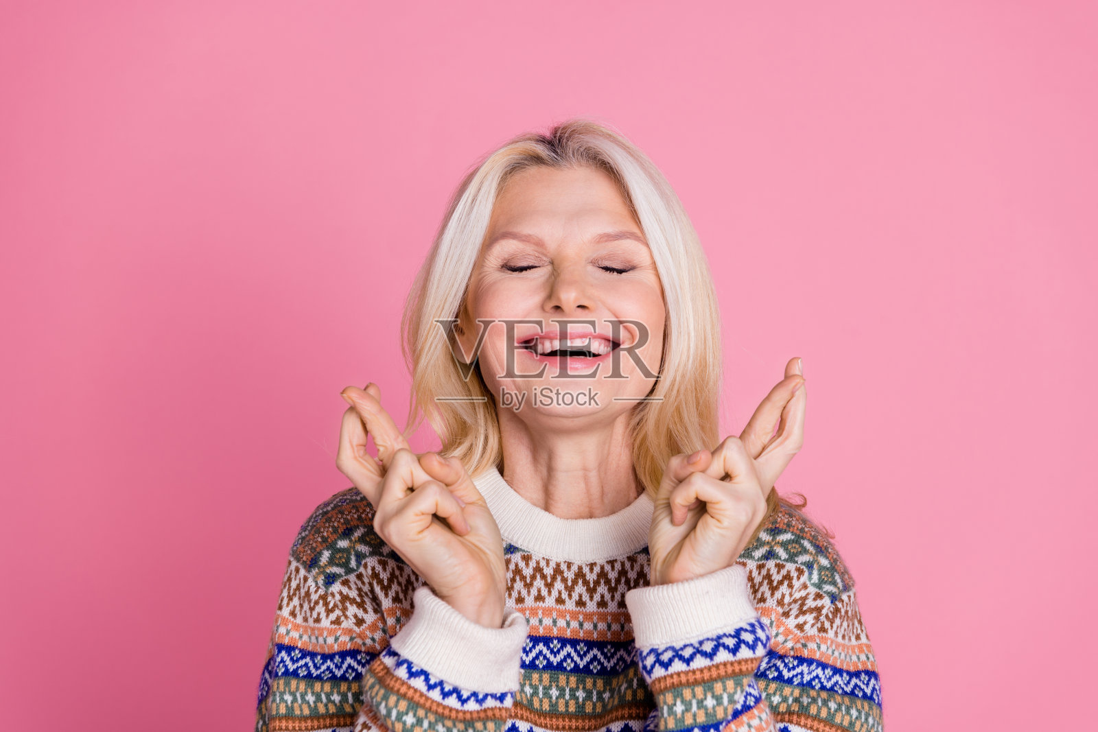 一位和蔼可亲的老年女士闭着眼睛,双手交叉,穿着毛衣,背景为粉色。照片摄影图片