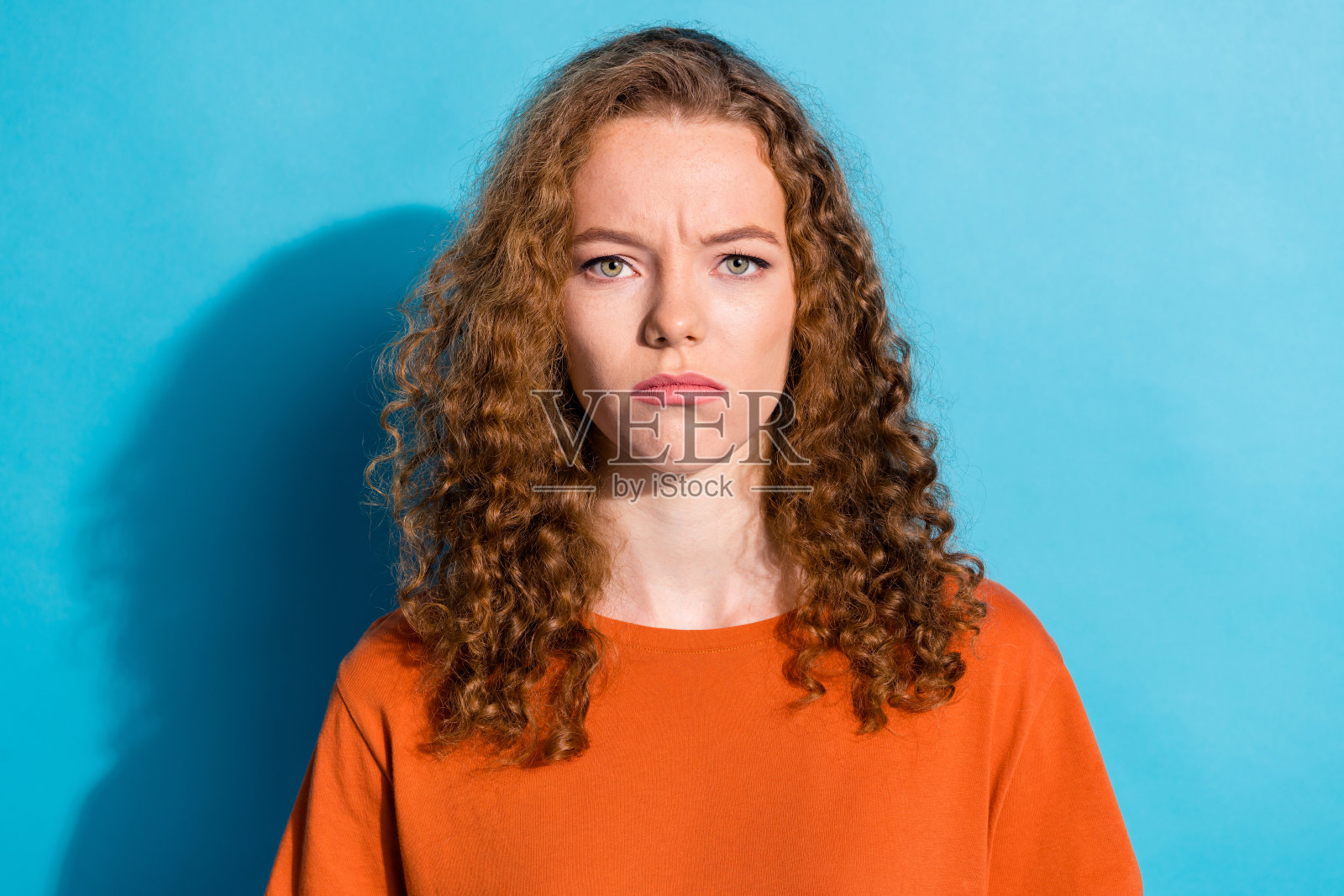 青春美丽的女孩,面露搞笑而不悦的表情,波浪卷发,穿着橙色T恤,背景为蓝色。照片摄影图片