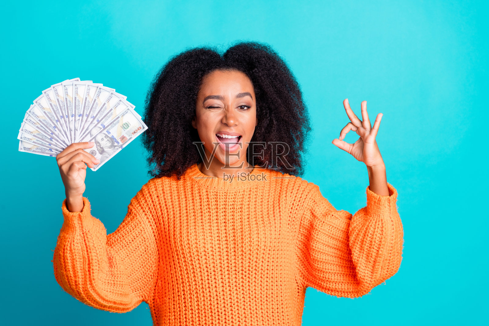 漂亮年轻女孩手持钞票扇,眨眼做出“好的”手势,穿着时尚的橙色针织服装,背景为水绿色。照片摄影图片