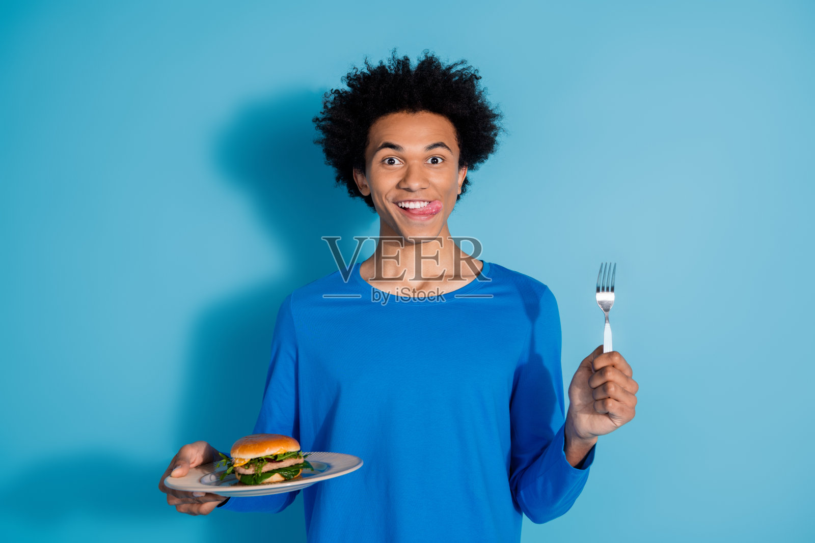 年轻帅气的男士在蓝色背景下吃汉堡,手持叉子,舔着嘴唇,穿着时尚的服装。照片摄影图片