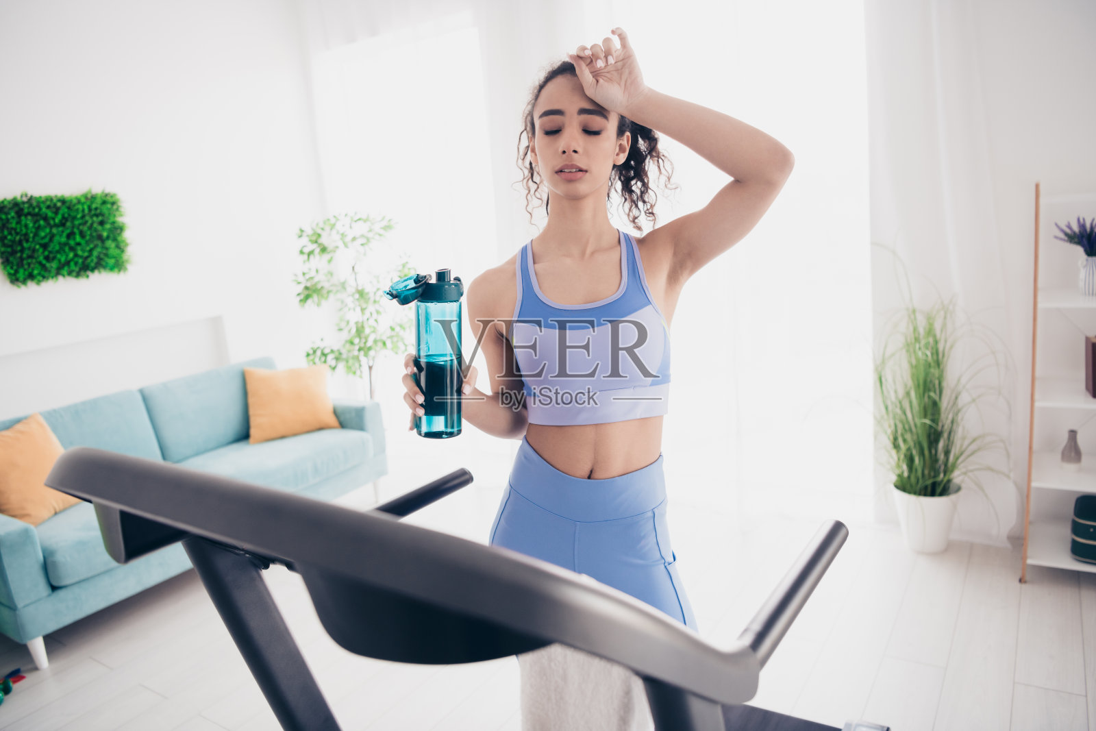 年轻女性在现代公寓内的跑步机上锻炼,手中拿着清爽饮料,保持健康和专注。照片摄影图片