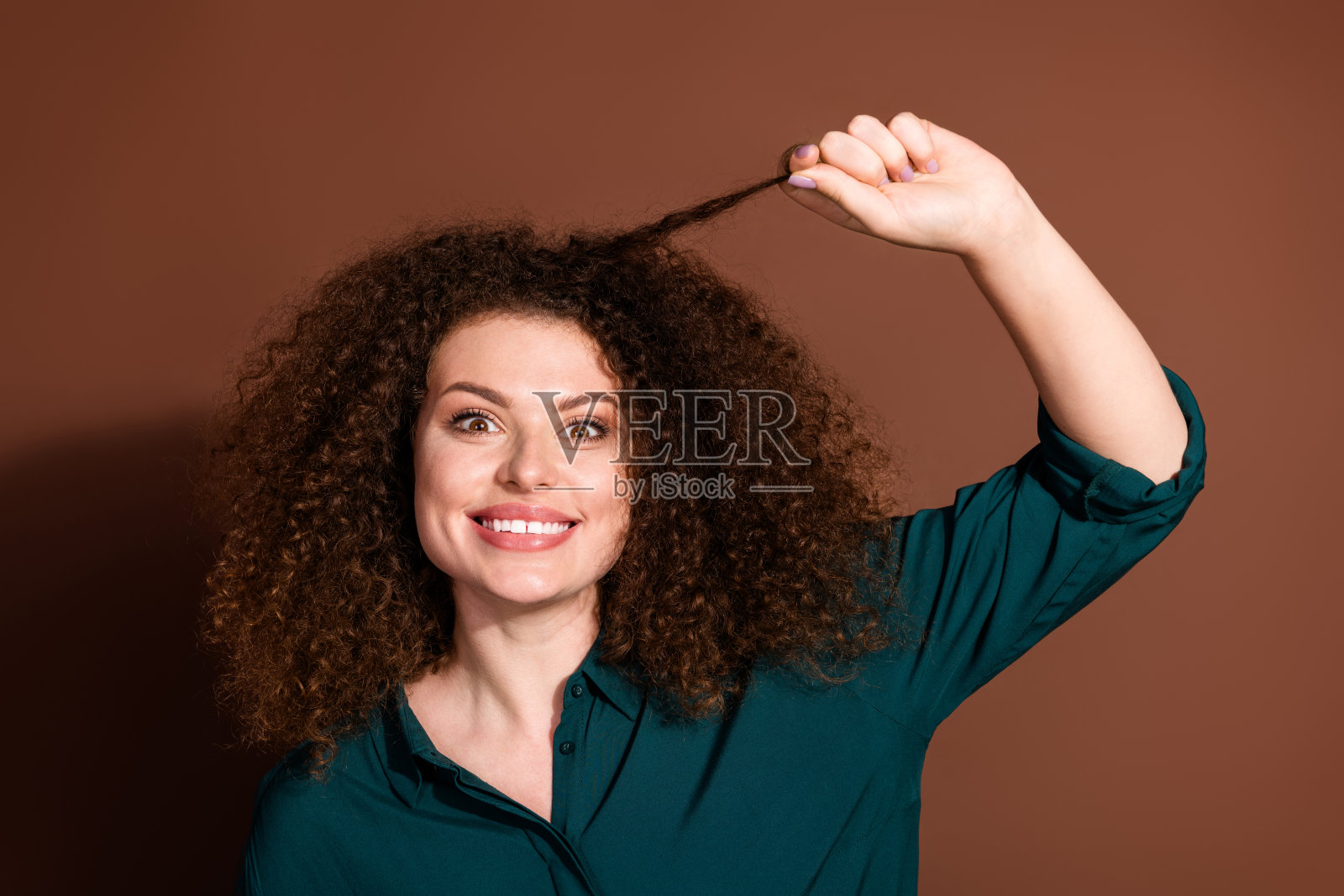 穿着绿色衬衫的快乐年轻女性,卷发,背景为棕色,灿烂微笑。照片摄影图片