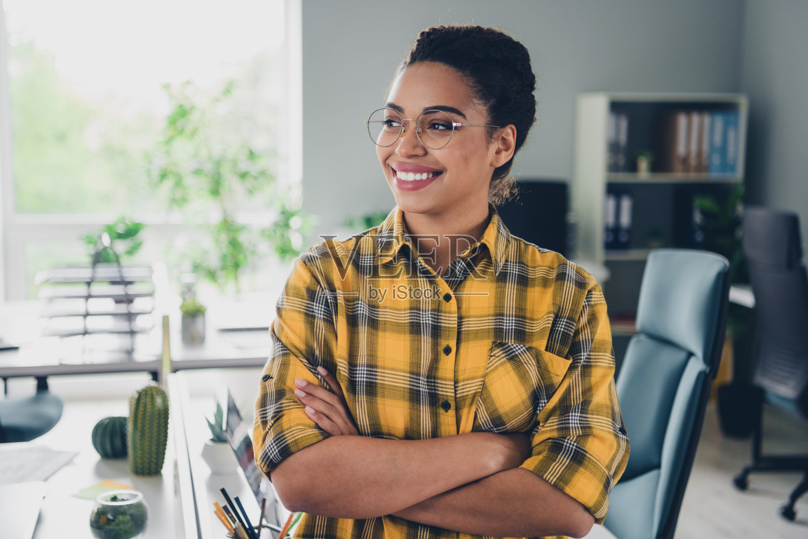 穿着黄色格子衬衫的可爱年轻女士双手交叉,自信地看着空旷的空间,坐在舒适的初创公司办公室内。照片摄影图片