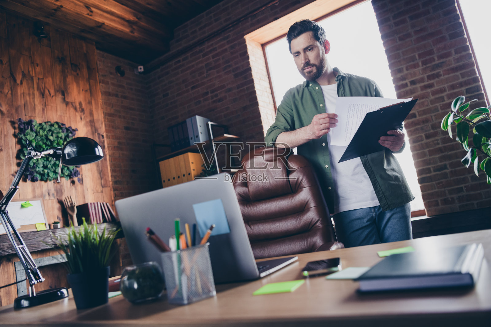 一位吸引人的年轻男子在现代办公室阁楼内的舒适工作空间中,穿着绿色衬衫,手持文件夹。照片摄影图片