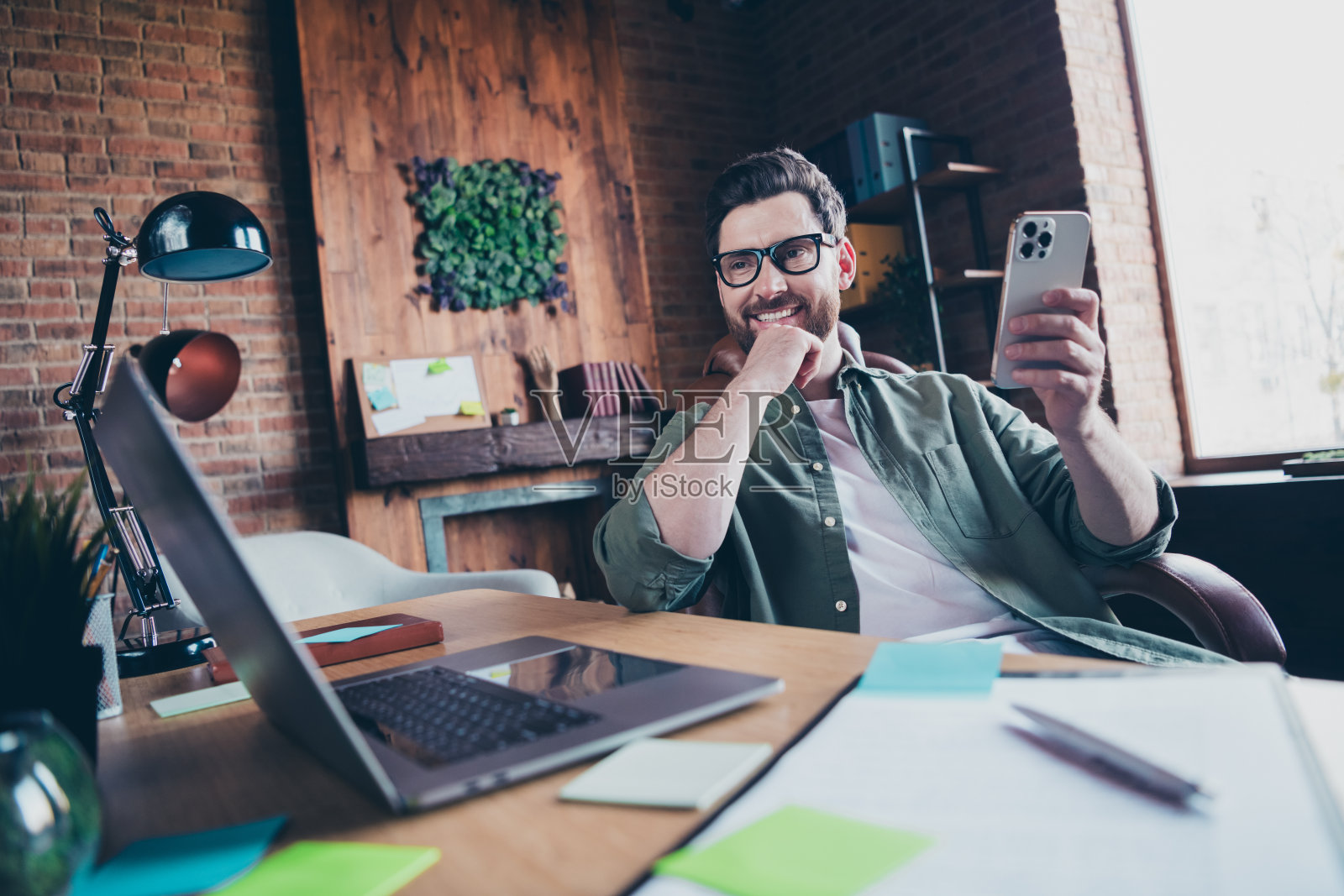 一位吸引人的年轻男子在现代办公室阁楼内的工作空间里,穿着绿色衬衫,舒适地使用着手持设备。照片摄影图片