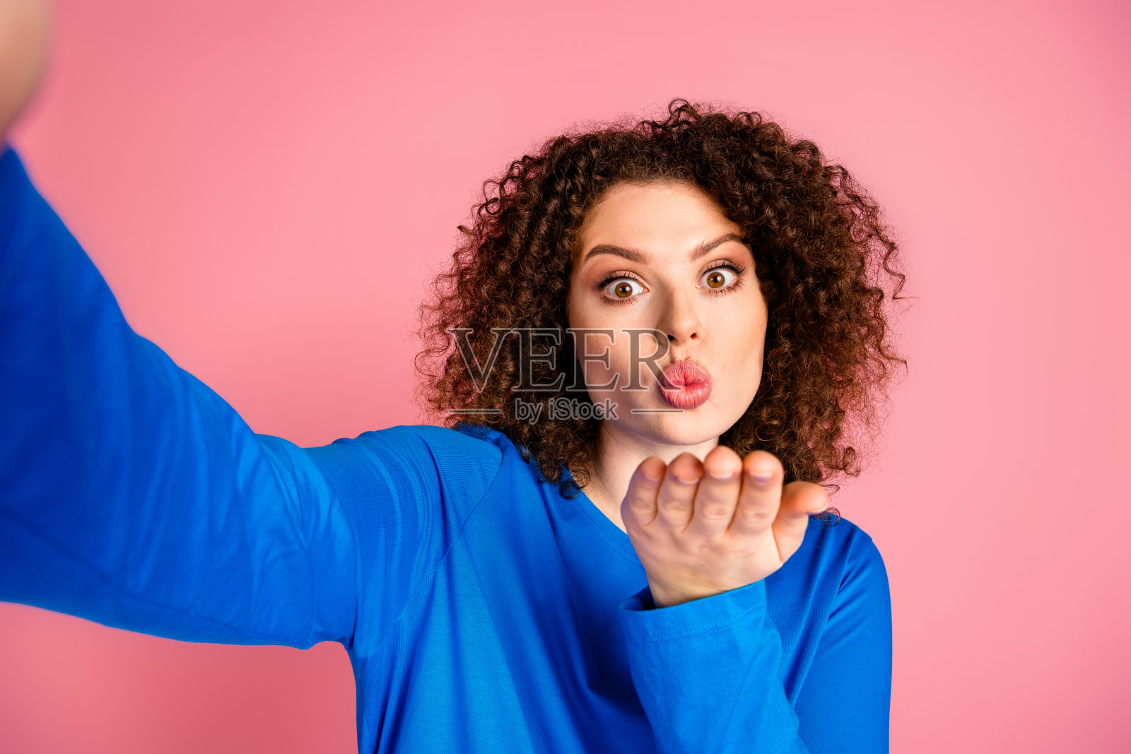 年轻女性穿着休闲蓝色卫衣,卷发吹吻,背景为粉色。照片摄影图片