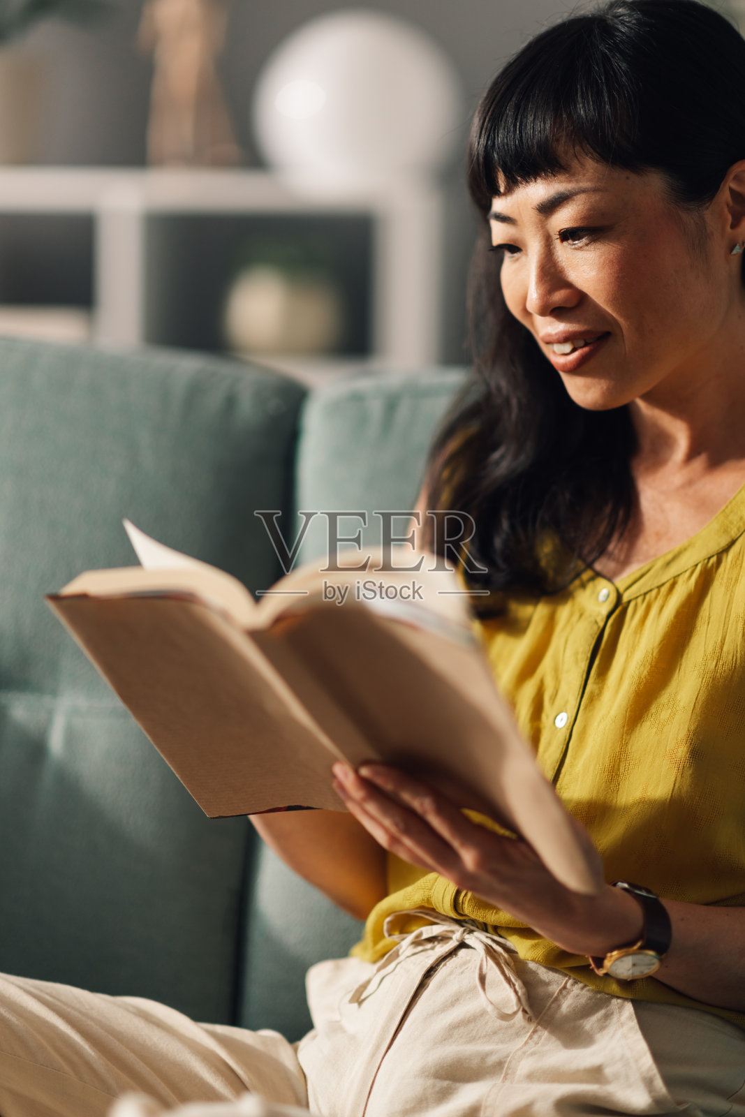 亚洲女性坐在舒适的绿色沙发上,沉浸在书本中。照片摄影图片