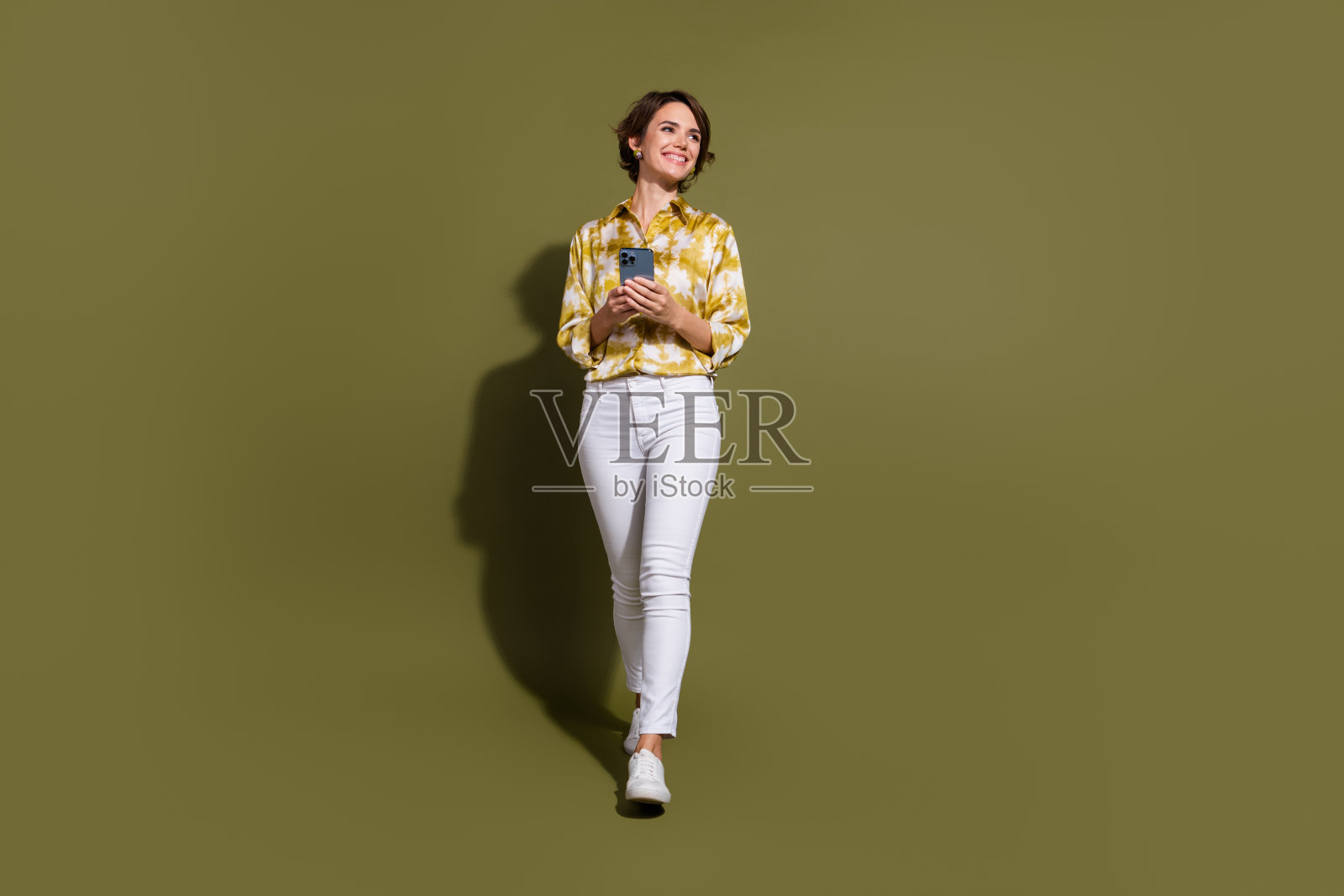 年轻职业女性在卡其色背景下,穿着时尚的上衣,手持手机,自信地微笑。照片摄影图片