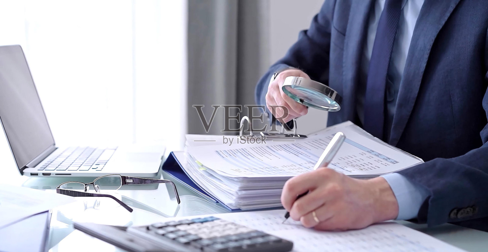 商人或审计师在使用计算器和放大镜分析财务文件时写笔记。审计和税务概念。照片摄影图片