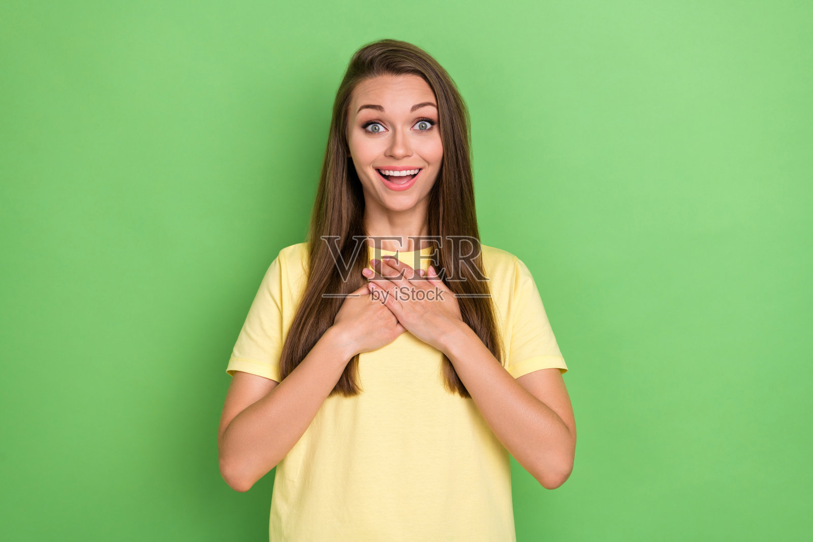 可爱的积极女孩穿着黄色T恤,双手放在胸前,背景为绿色,展现出难以置信的胜利。照片摄影图片