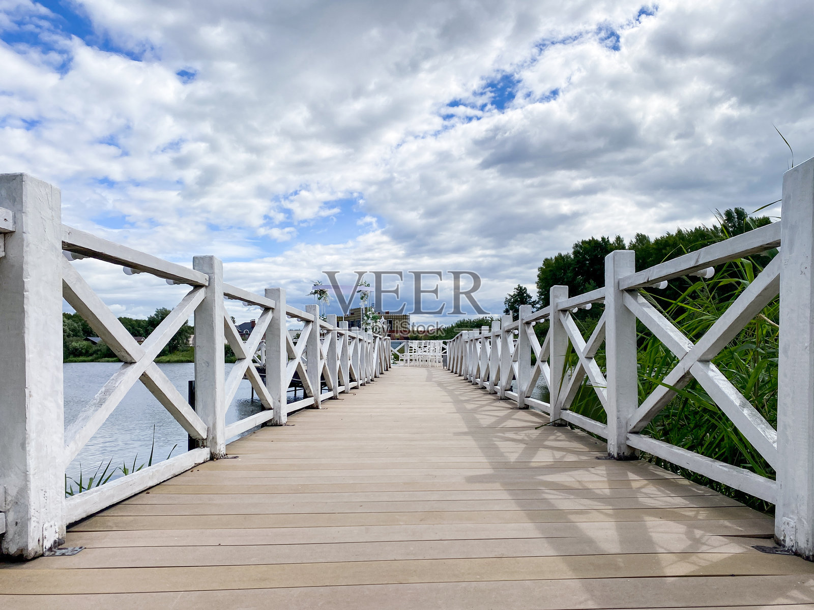 一座如画的木桥横跨在宁静的水道上,周围被生机勃勃的绿树环绕,壮观的多云天空为这一幕增添了戏剧性。照片摄影图片