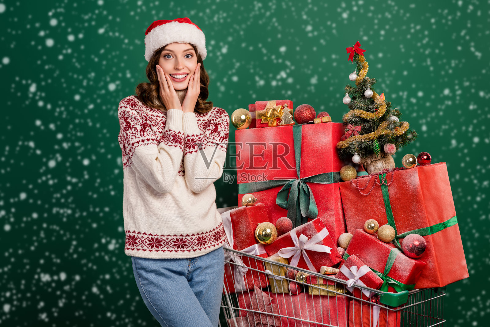 兴奋的积极女孩双手捧着脸颊,堆满礼品盒的购物车,背景为绿色。照片摄影图片
