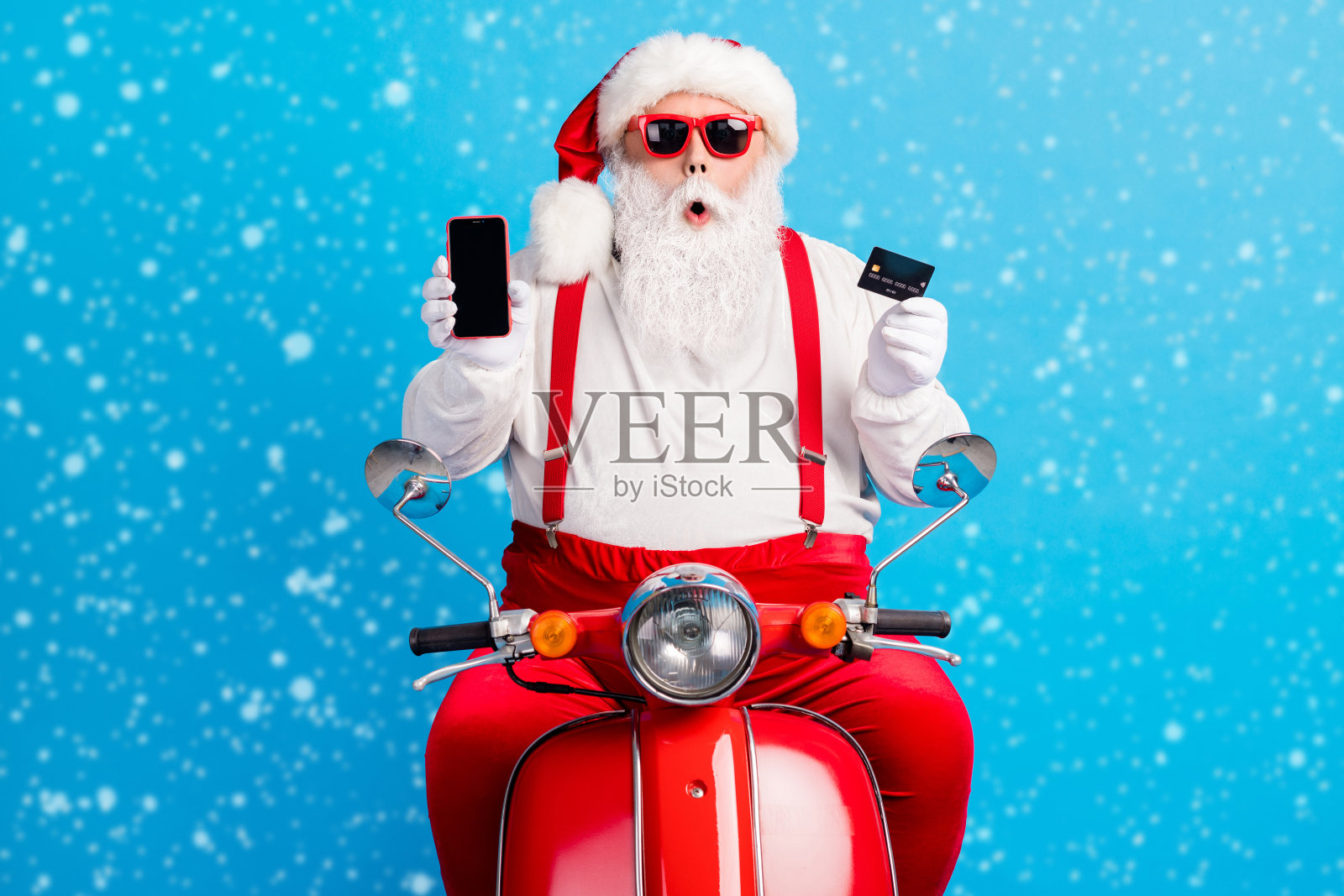 穿着圣诞服装、吊带裤、太阳镜和白衬衫的祖父,留着灰色胡须,骑着复古摩托车,手持借记卡,手机屏幕空白,背景为蓝色。照片摄影图片