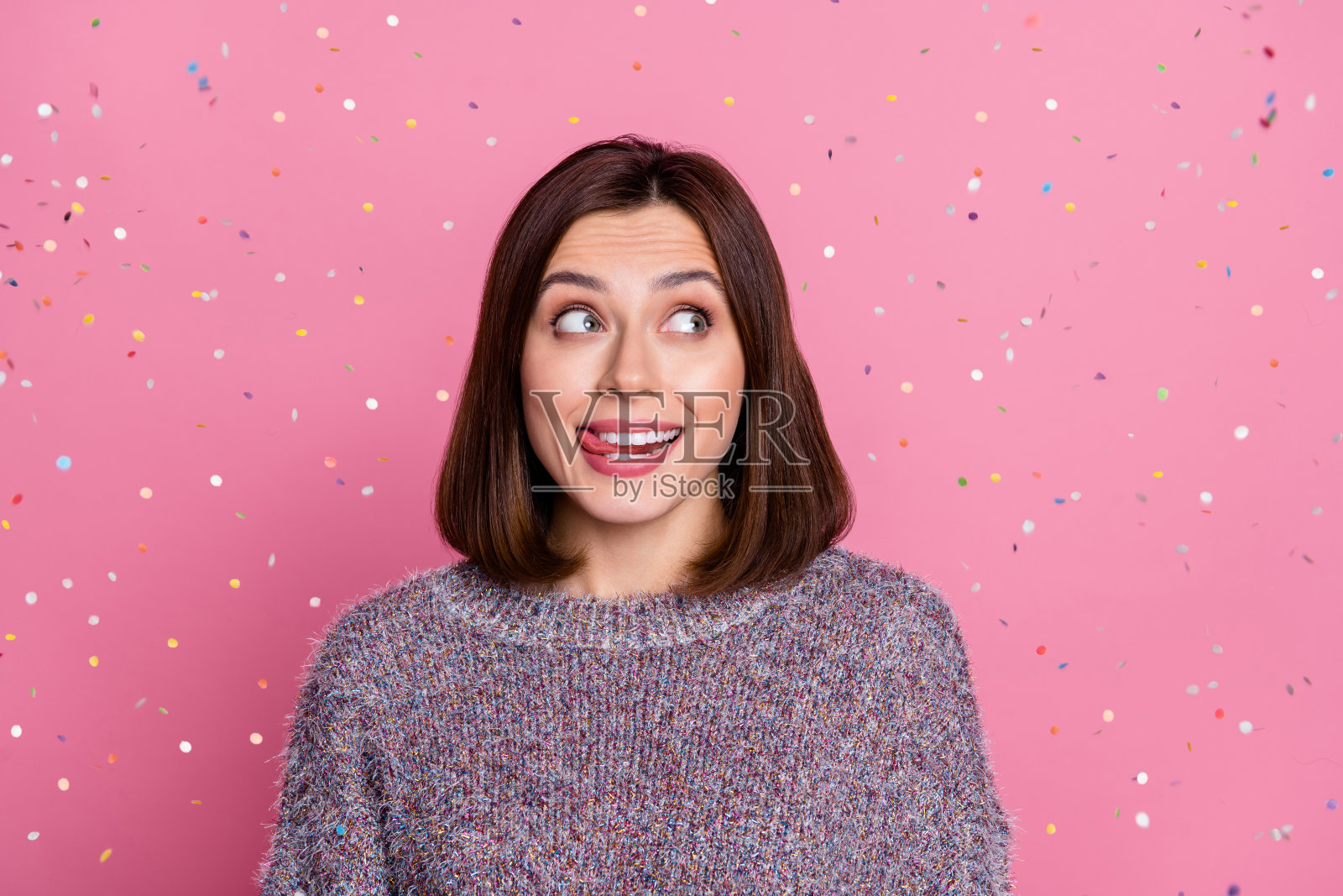 一位迷人的棕发时尚快乐女孩在粉色柔和背景下飞舞着五彩纸屑,舔着嘴唇。照片摄影图片