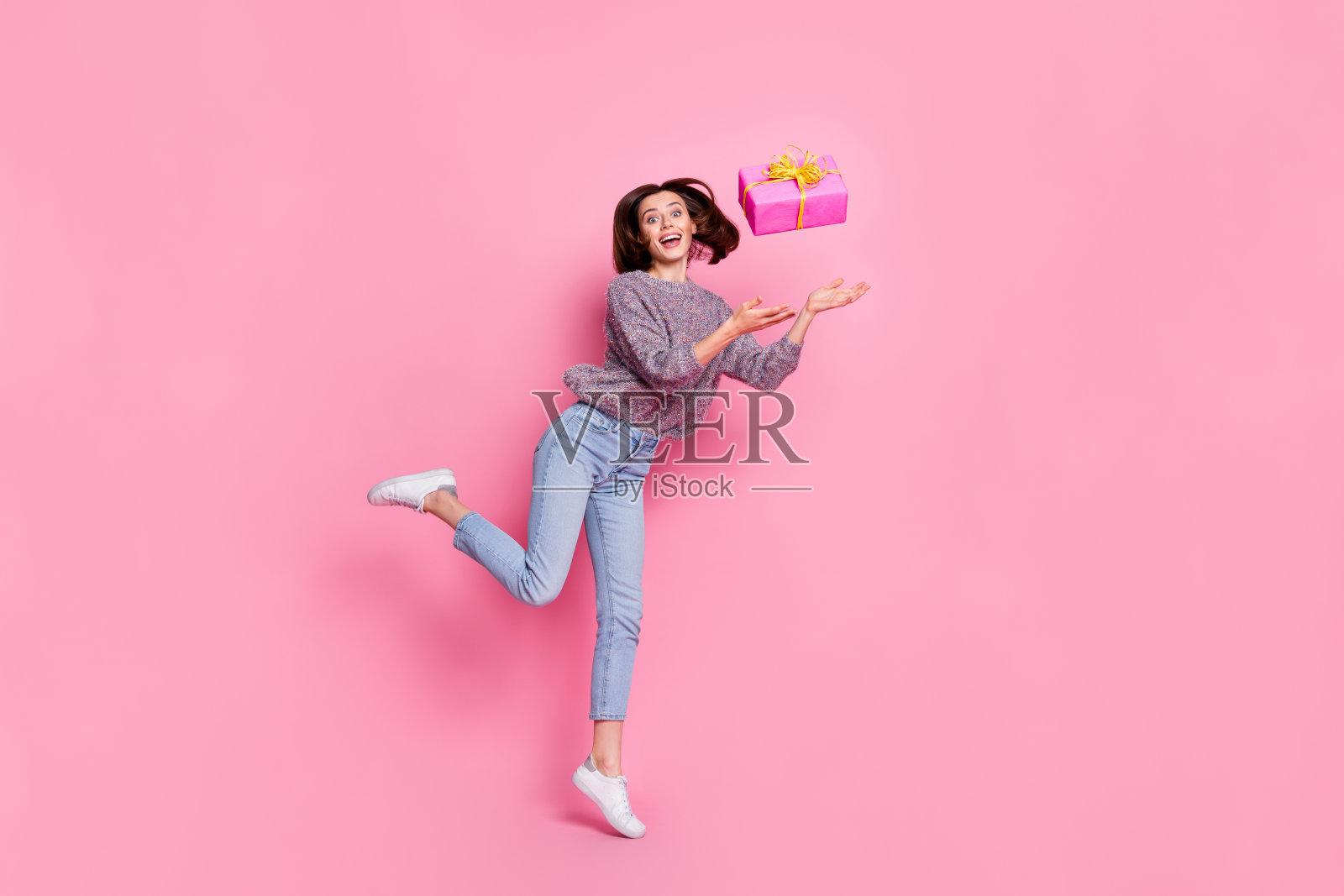 穿着毛衣的可爱惊讶女士在粉色背景上跳起扔礼物。照片摄影图片