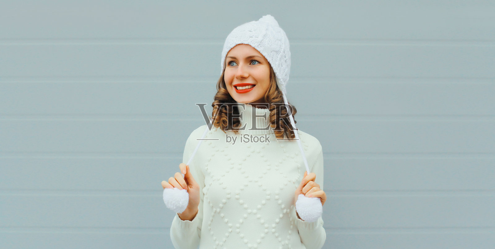 冬季肖像,快乐的年轻女性微笑着,戴着白色帽子和毛衣,目光投向灰色背景。照片摄影图片