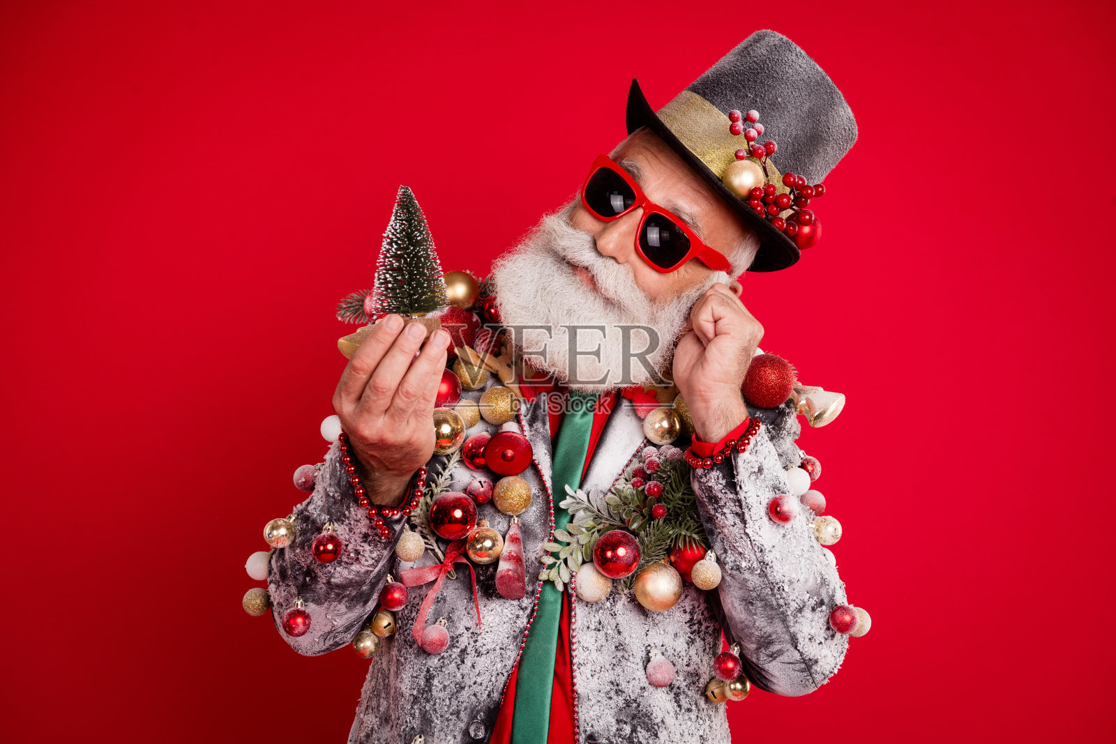 穿着优雅时尚西装、佩戴夏季眼镜和高顶帽的奇特奢华男士,手持圣诞玩具,背景为红色。照片摄影图片