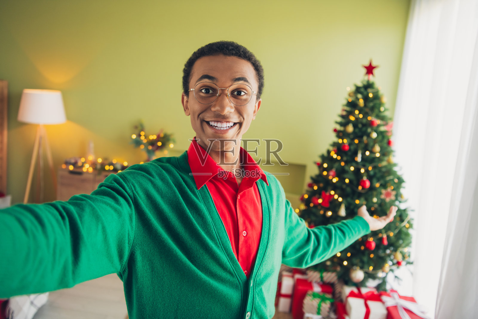年轻男子穿着绿色圣诞服装,愉快地在室内拍摄自拍,背景是温馨的圣诞派对装饰和树木。照片摄影图片