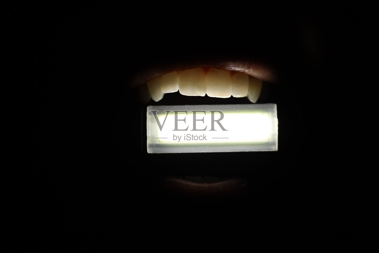 女孩在完全黑暗中用牙齿夹着一只小手电筒,照亮周围。照片摄影图片