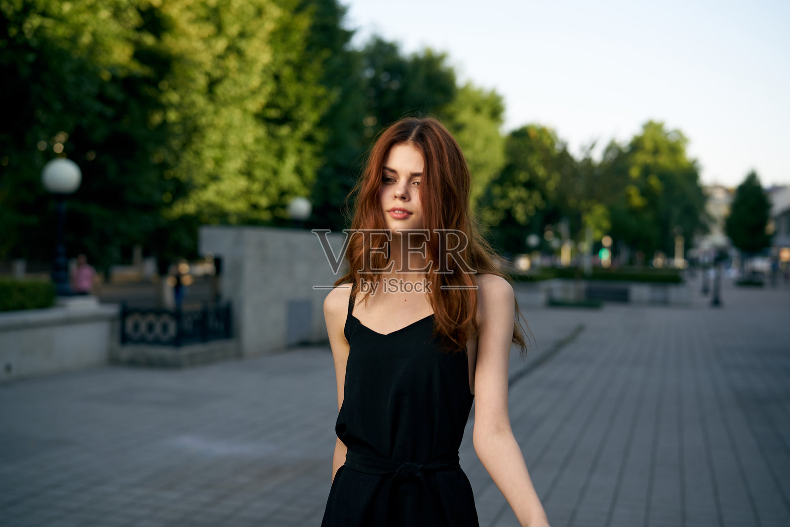 时尚女性在城市街道上,身穿黑色连衣裙,佩戴帽子和眼镜,化着妆,手腕上戴着手表,享受新鲜空气。照片摄影图片