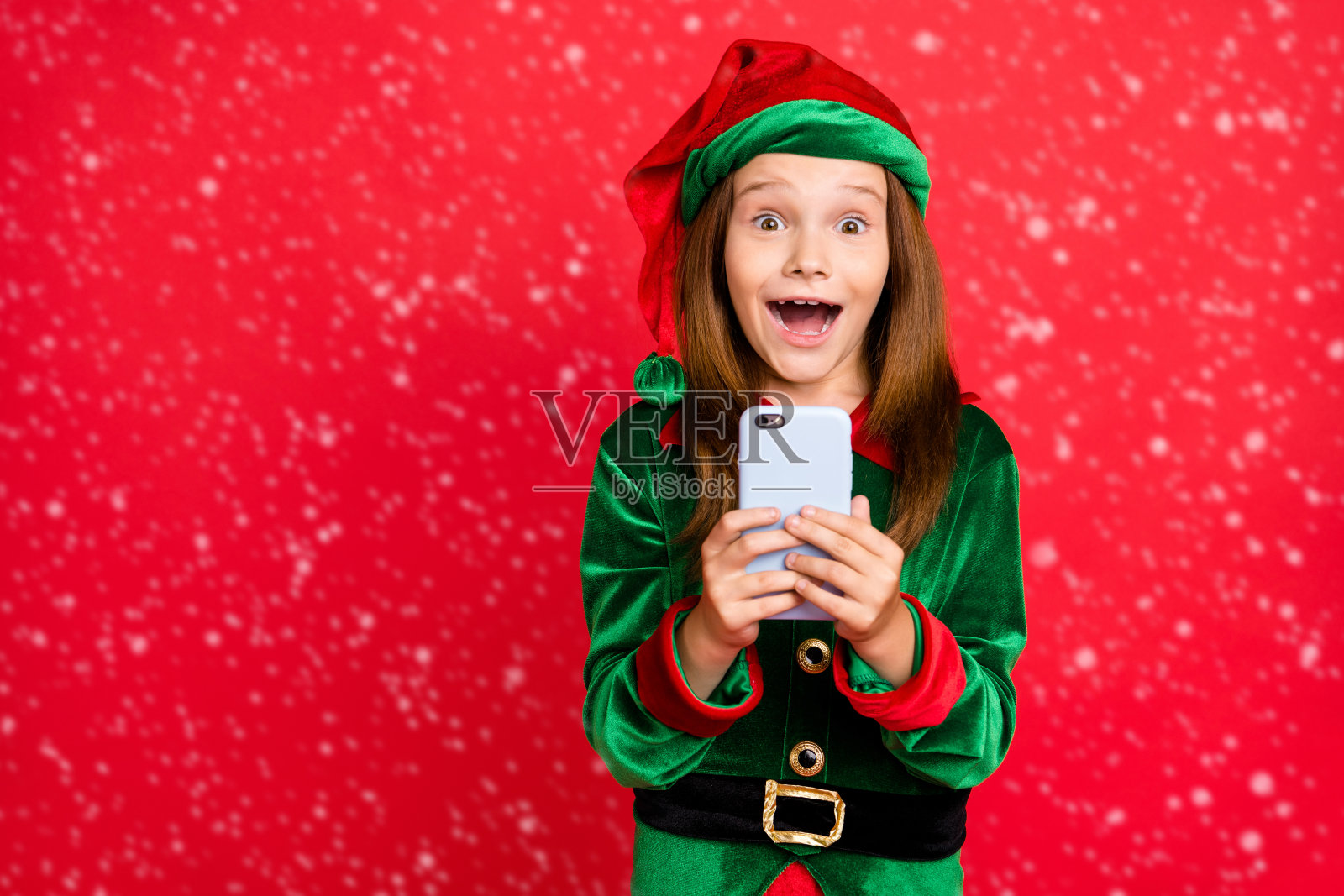 惊讶的精灵肖像,戴着绿色帽子,长长的红发,惊呼“哇,天哪”,使用智能手机获取点赞,评论穿着精灵服装,背景为红色。照片摄影图片