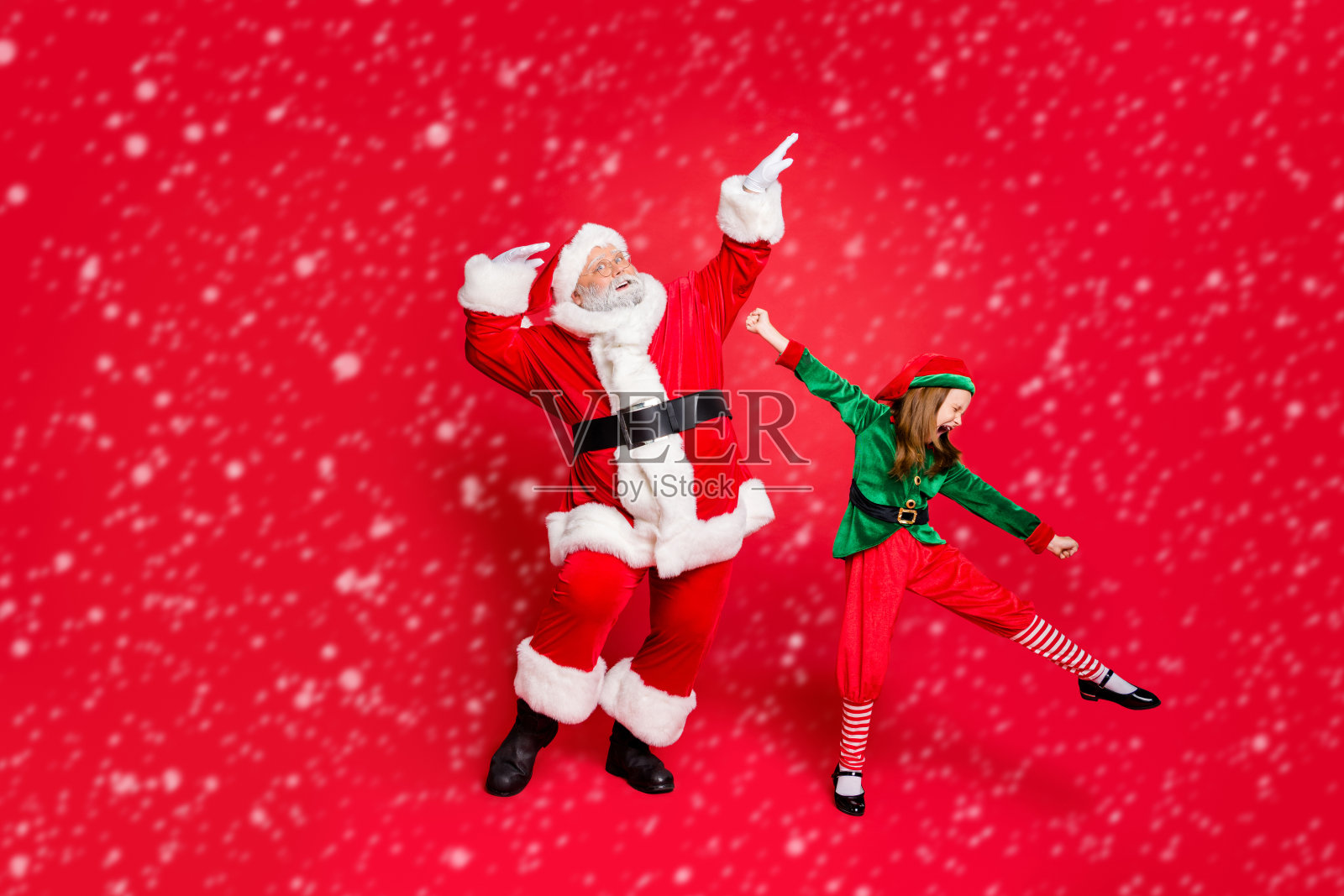 兴奋的圣诞老人和戴眼镜的精灵在红色背景上穿着亮丽的服装跳舞和搞笑的全身照片照片摄影图片