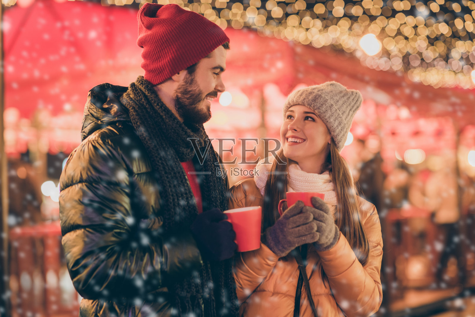 一对积极向上的情侣在户外的圣诞灯光下,手持传统的蛋奶酒饮品杯,穿着季节性外套。照片摄影图片