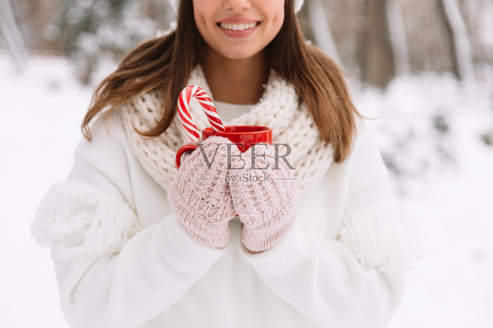 女人在公园里特写握着冬季杯子。女人戴着手套,手中拿着一个温暖的杯子,里面装着热可可、茶或咖啡,还有一根糖果手杖。冬季和圣诞节的概念。照片摄影图片