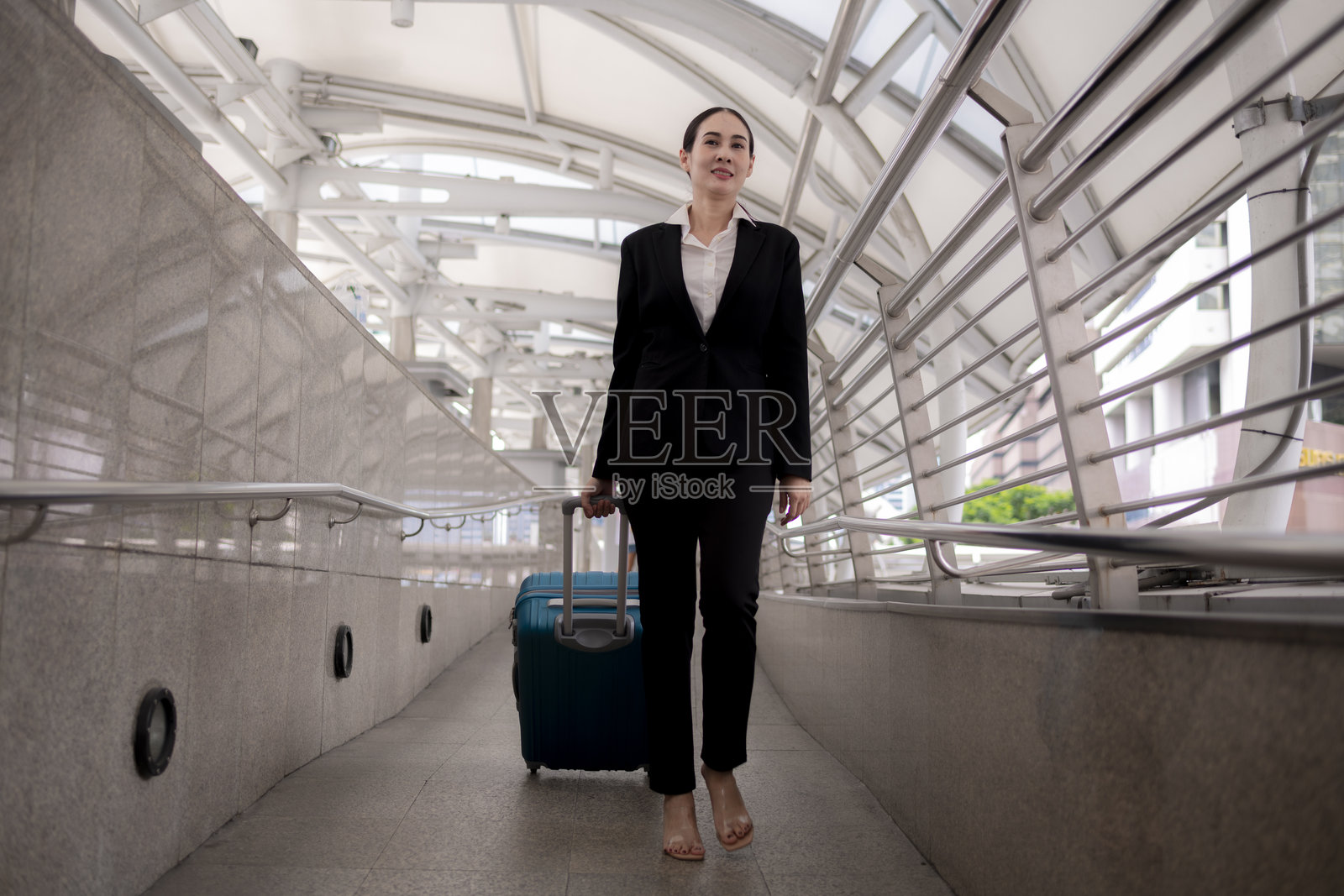 身穿西装的女商人站在公共交通或旅行中心,旁边有一个蓝色的拉杆行李箱,手中拿着平板或设备,正在查看。照片摄影图片