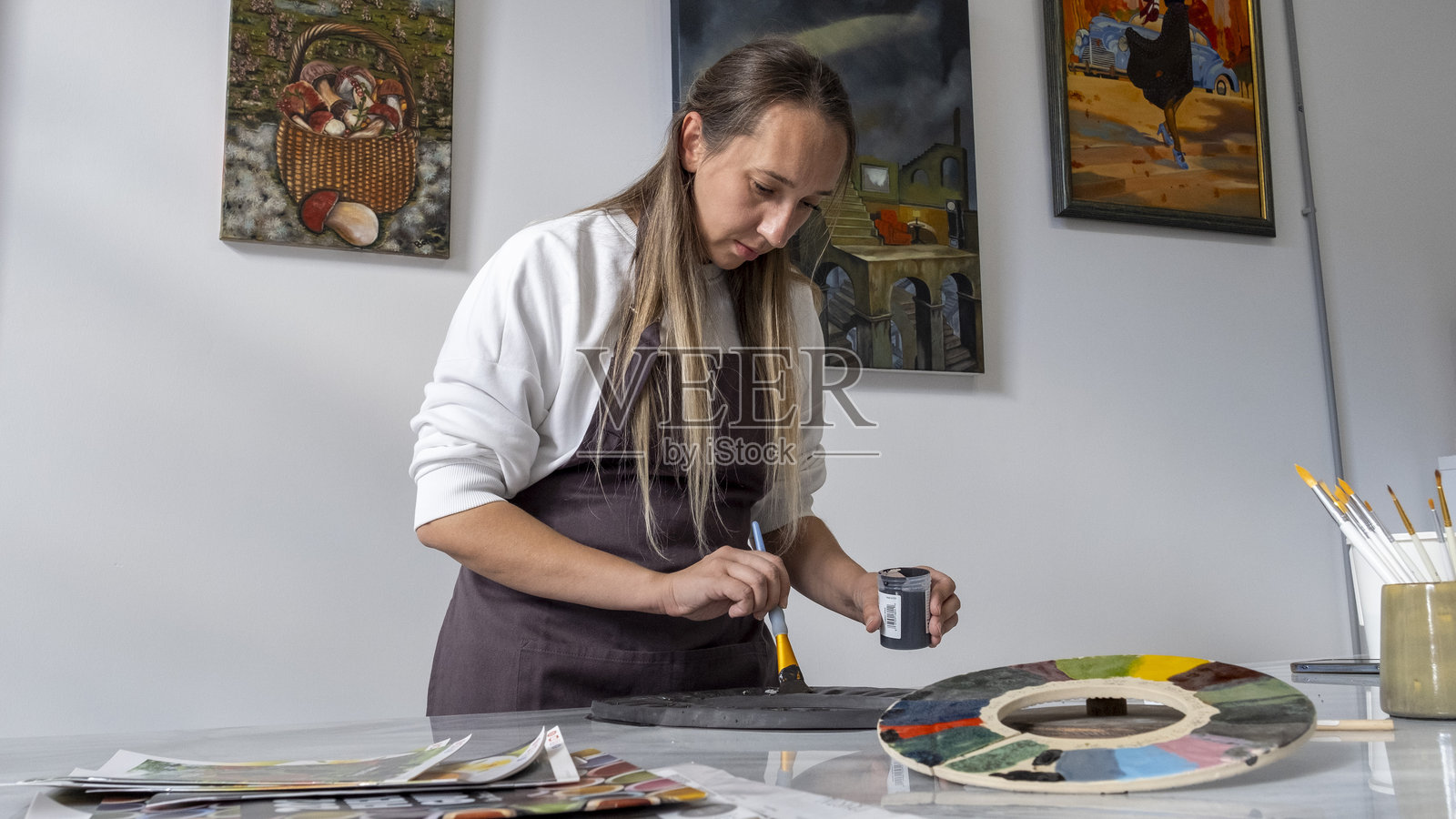 在一个艺术工作室,背景装饰着艺术画作,一位美丽的中年女性正在桌子上将一个陶土框架涂成黑色。照片摄影图片