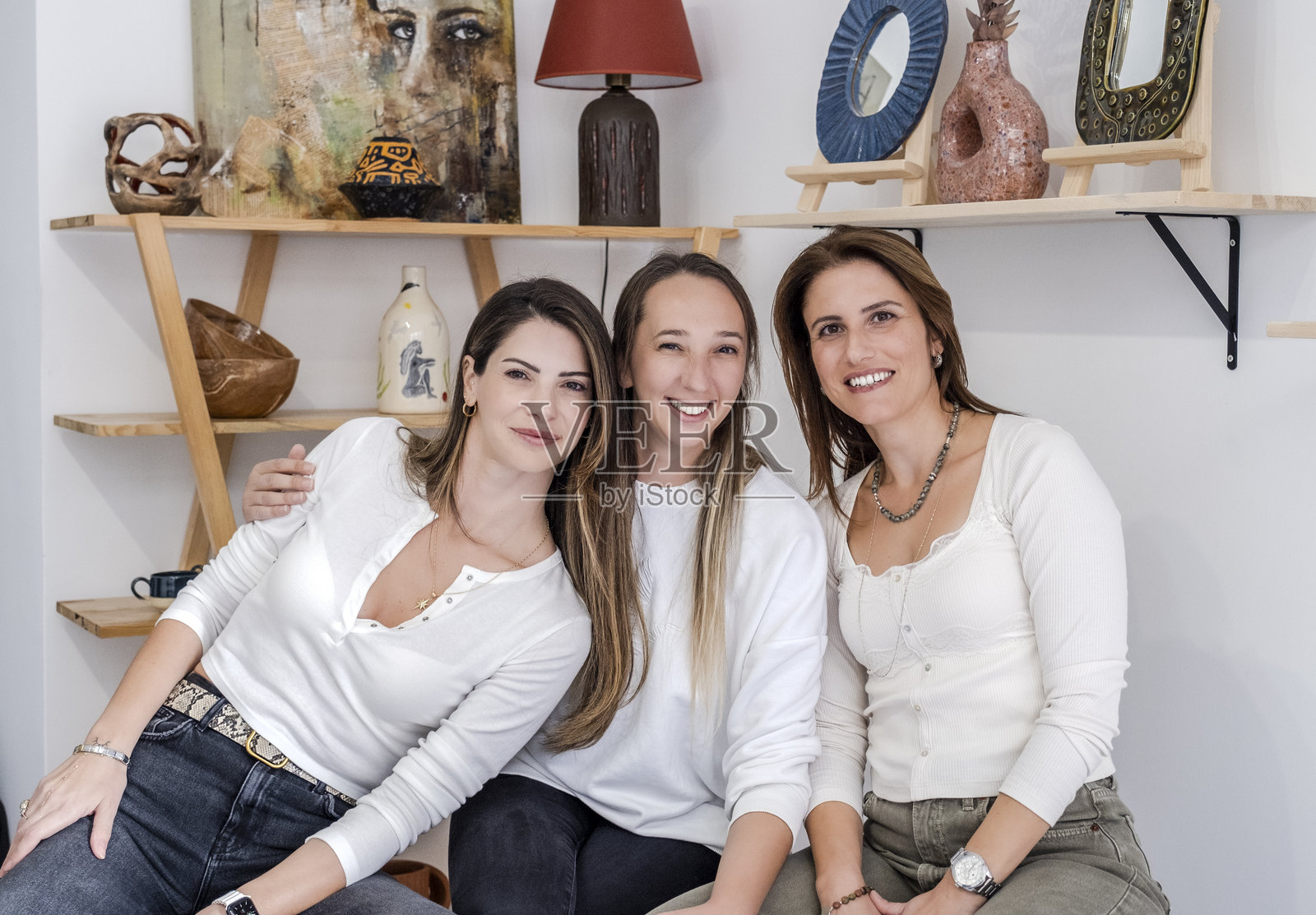 在一个艺术工作室,背景装饰着艺术画作,三位女性艺术家微笑着从前面的桌子上看向镜头。照片摄影图片