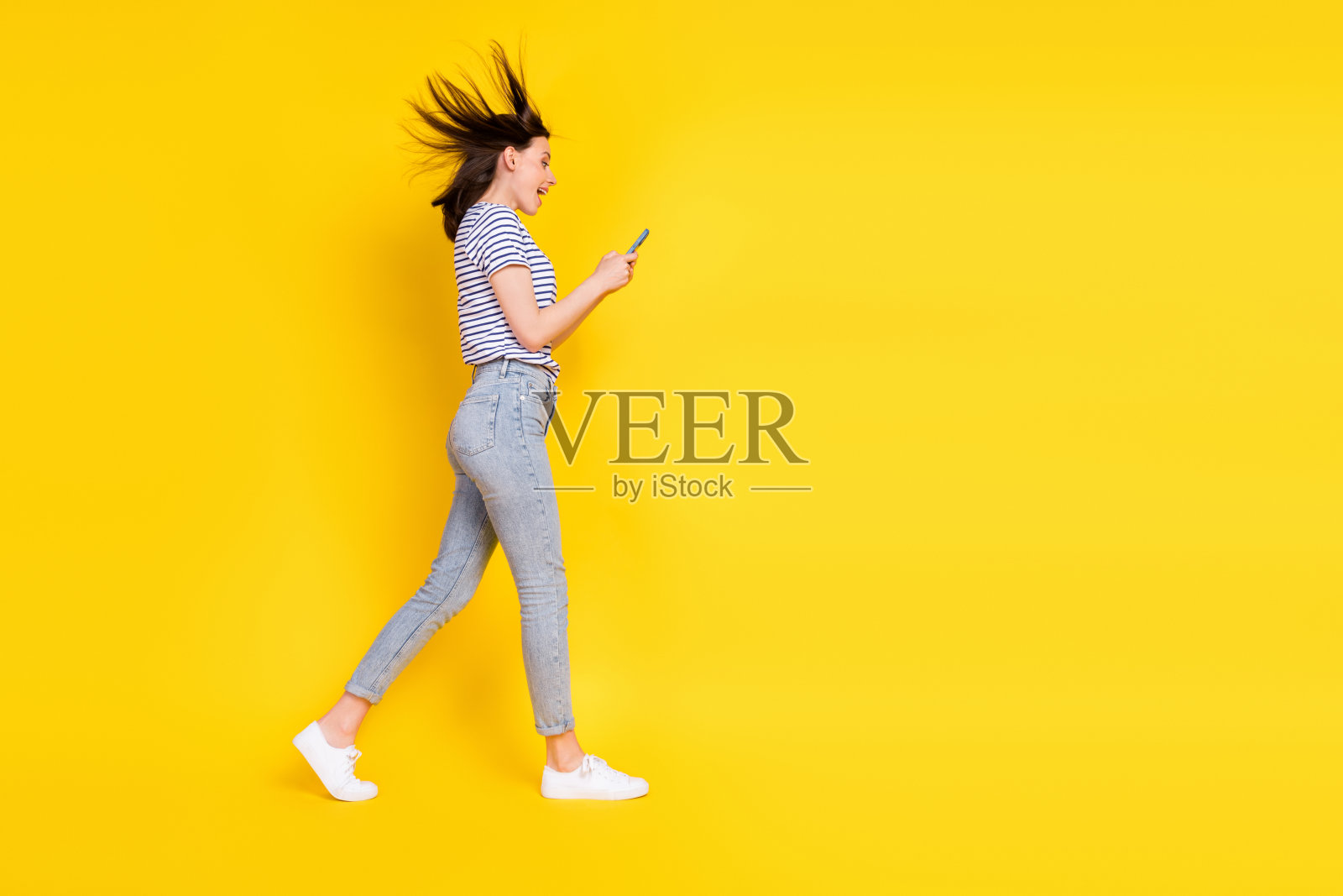 全身侧面照,时尚女性沉迷于手机设备,站在空旷的黄色背景前。照片摄影图片