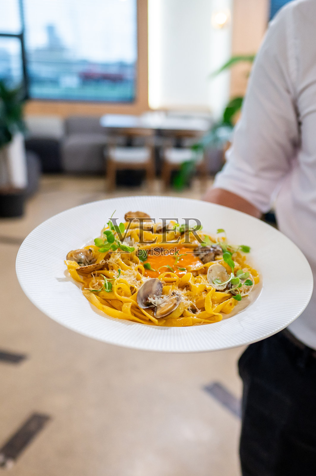 一位服务员在餐厅环境中端着一盘带有蛤蜊和香草的意大利面特写,展示了菜肴的鲜艳色彩和新鲜食材。非常适合烹饪和用餐主题。照片摄影图片