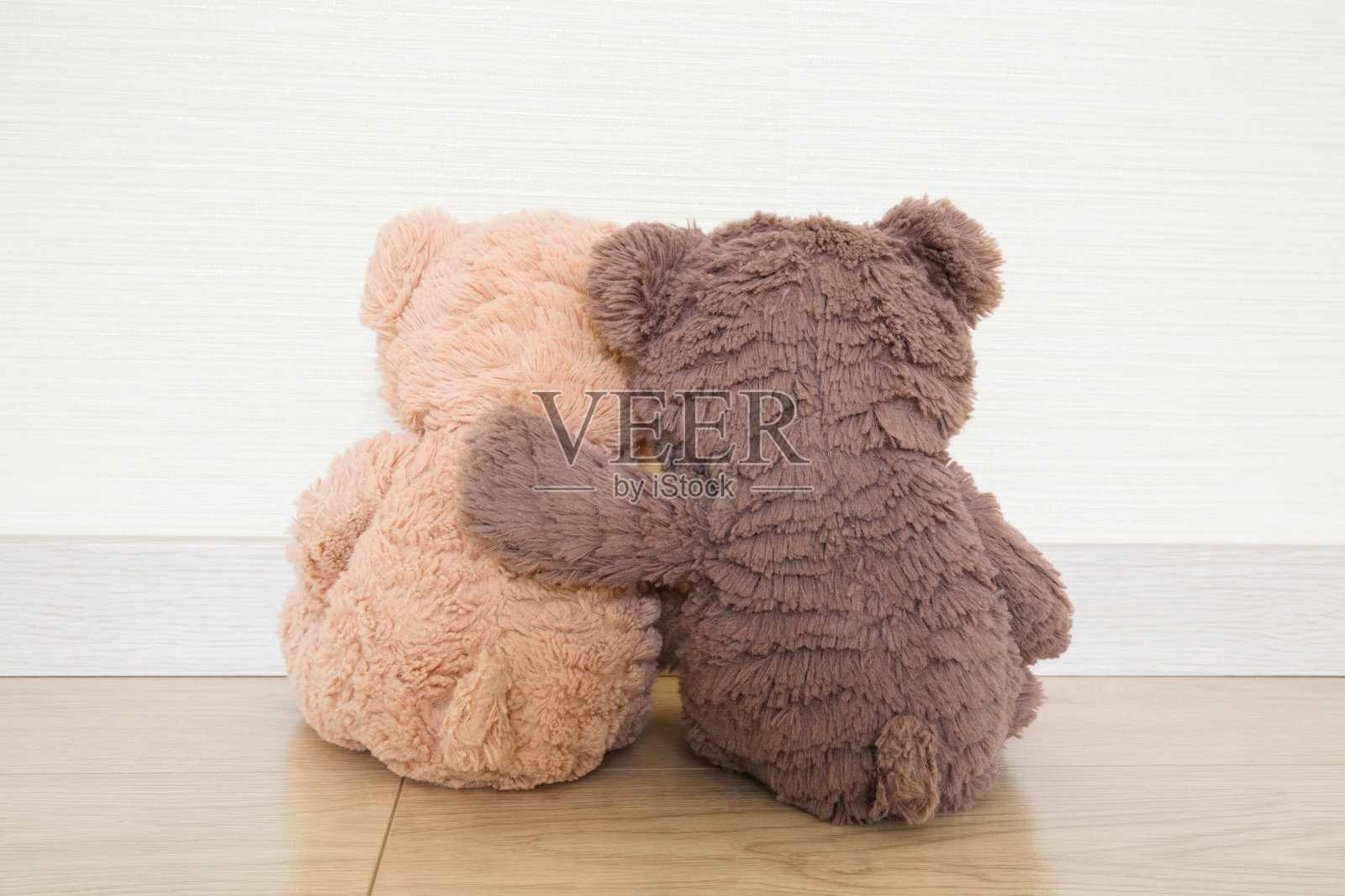 两只浅棕色和深棕色的软绒泰迪熊相互拥抱,坐在木地板上,背景是浅灰色的墙壁。特写。背面视角。照片摄影图片
